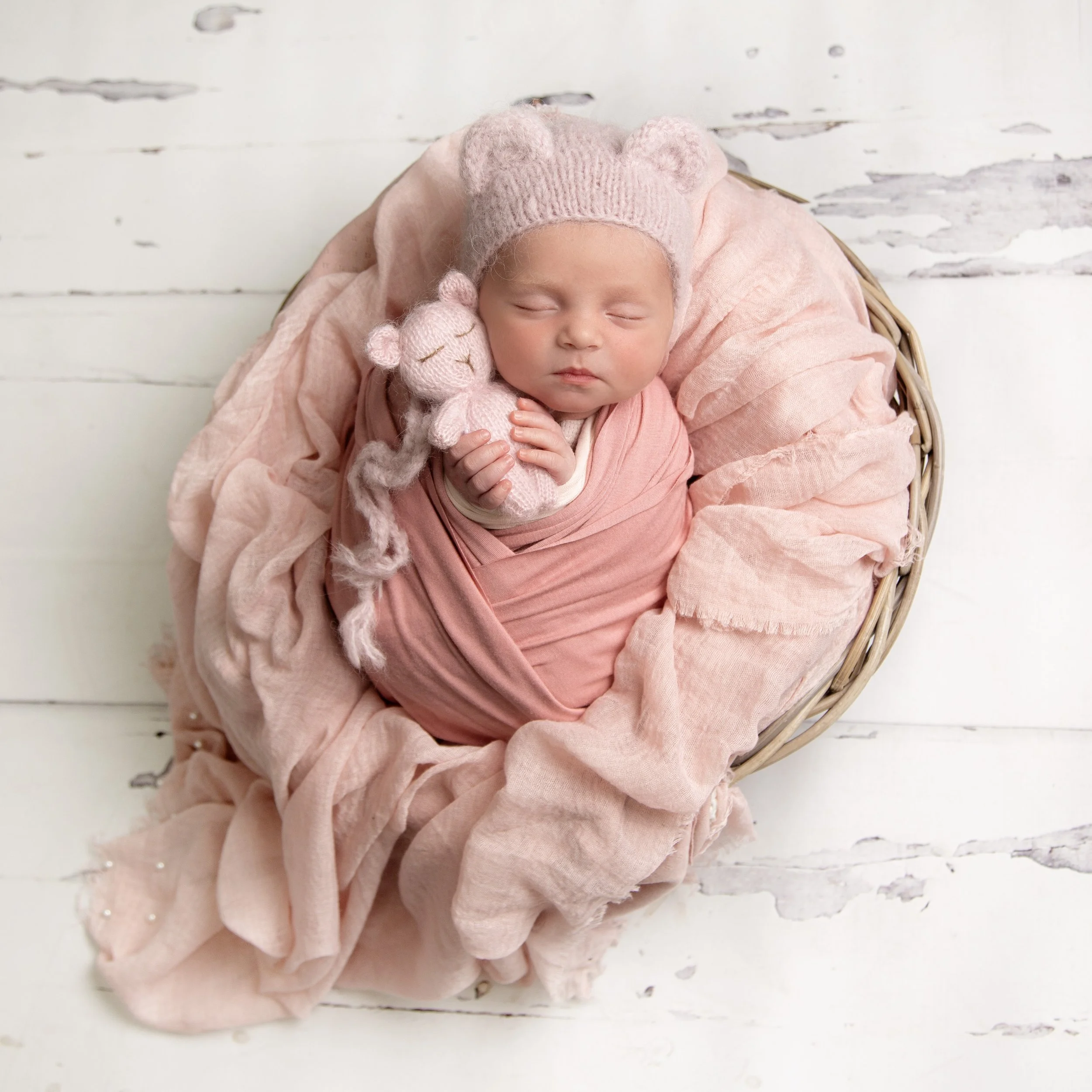 A sleeping baby wrapped in pink cloth, wearing a knitted pink bear hat, holding a small knitted bear toy, lying in a woven basket on a white wooden floor with distressed paint.