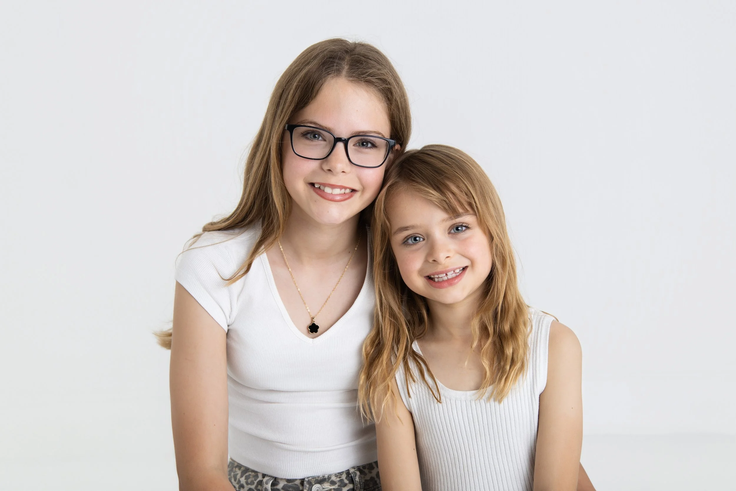 A young woman and a young girl smiling, sitting close together against a plain white background.