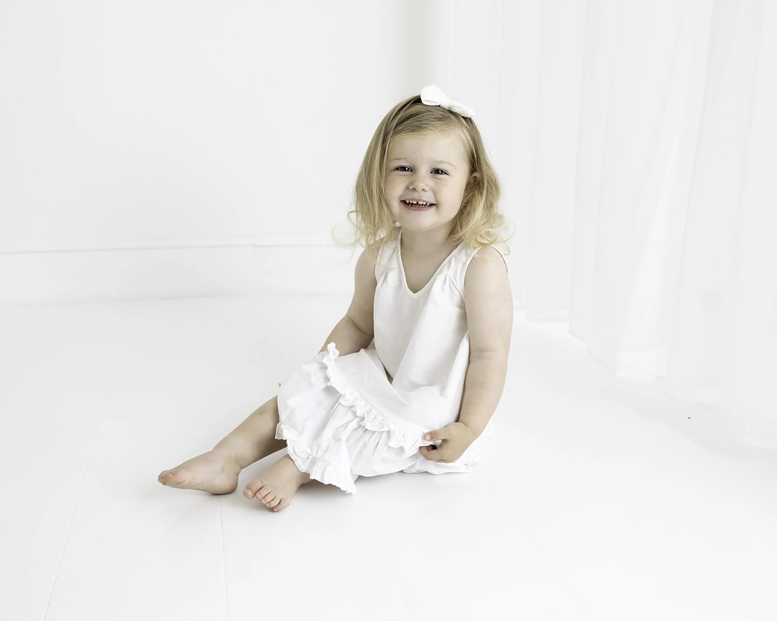 Young girl sitting on white floor, smiling, wearing a white dress and a white bow in her hair, with sheer white curtains in the background.