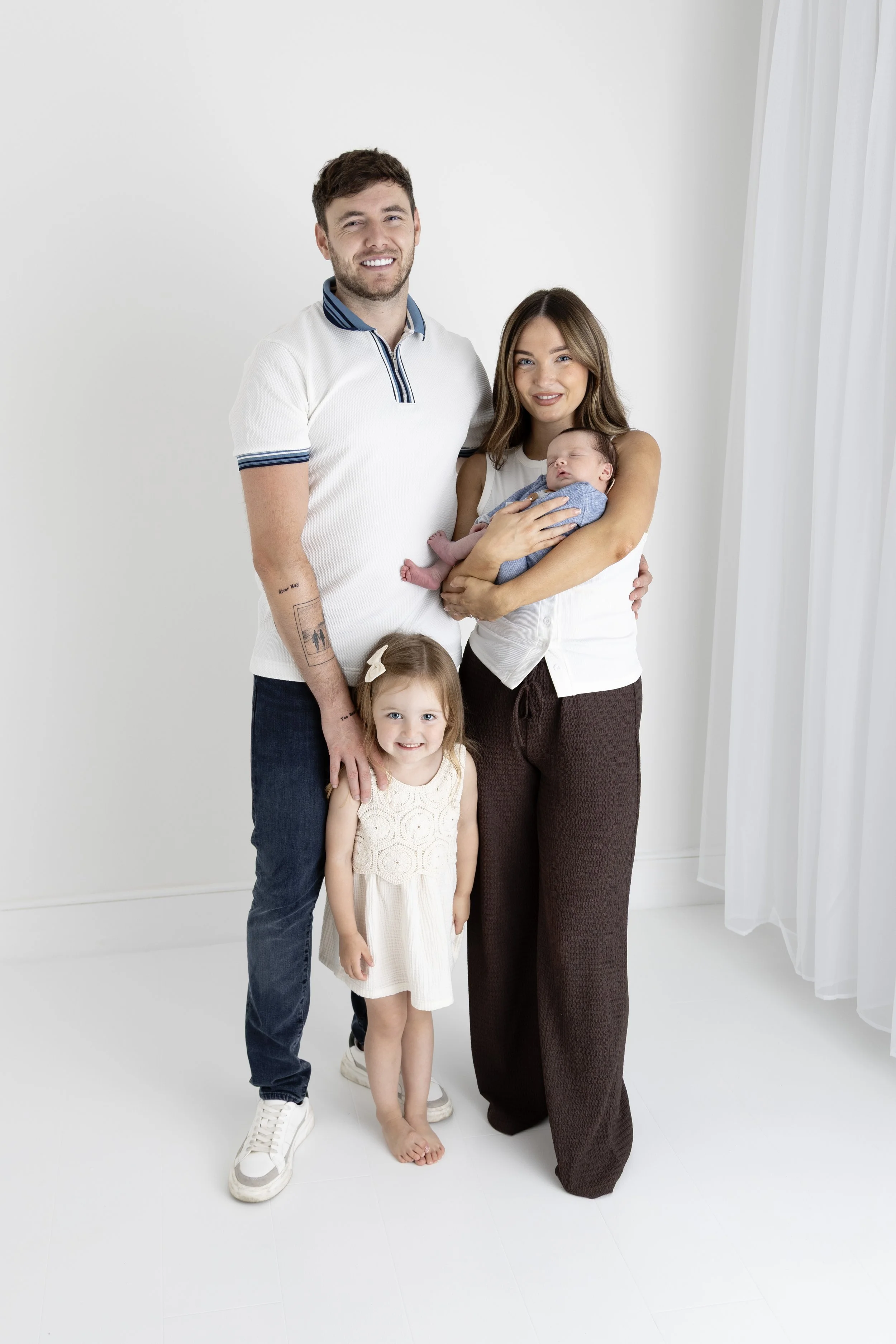 A family of four standing together in a white room with sheer white curtains. The mother is holding a sleeping baby, and a young girl is standing beside her, smiling. The father stands next to them, smiling.