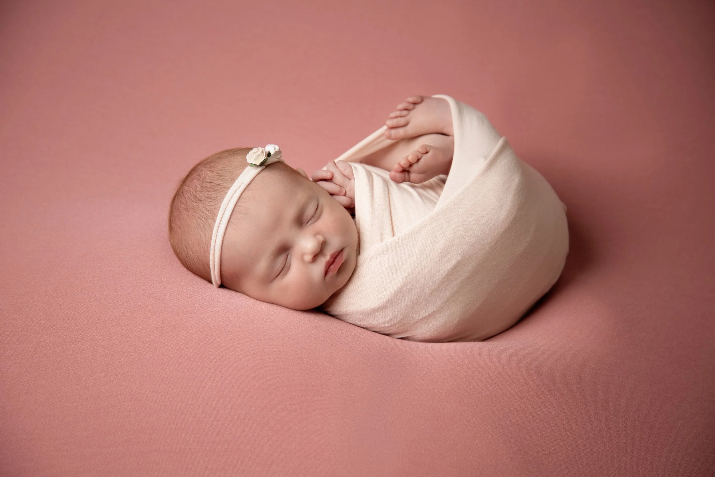 A sleeping newborn baby girl wrapped in a soft pink blanket, wearing a headband with a small flower, resting on a pink surface.