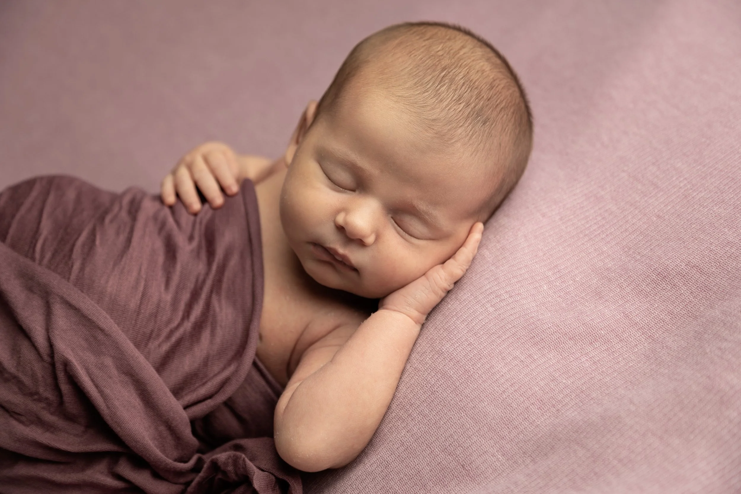 A sleeping Asian baby lying sideways on a pink surface, with head resting on one hand, wearing a maroon outfit.