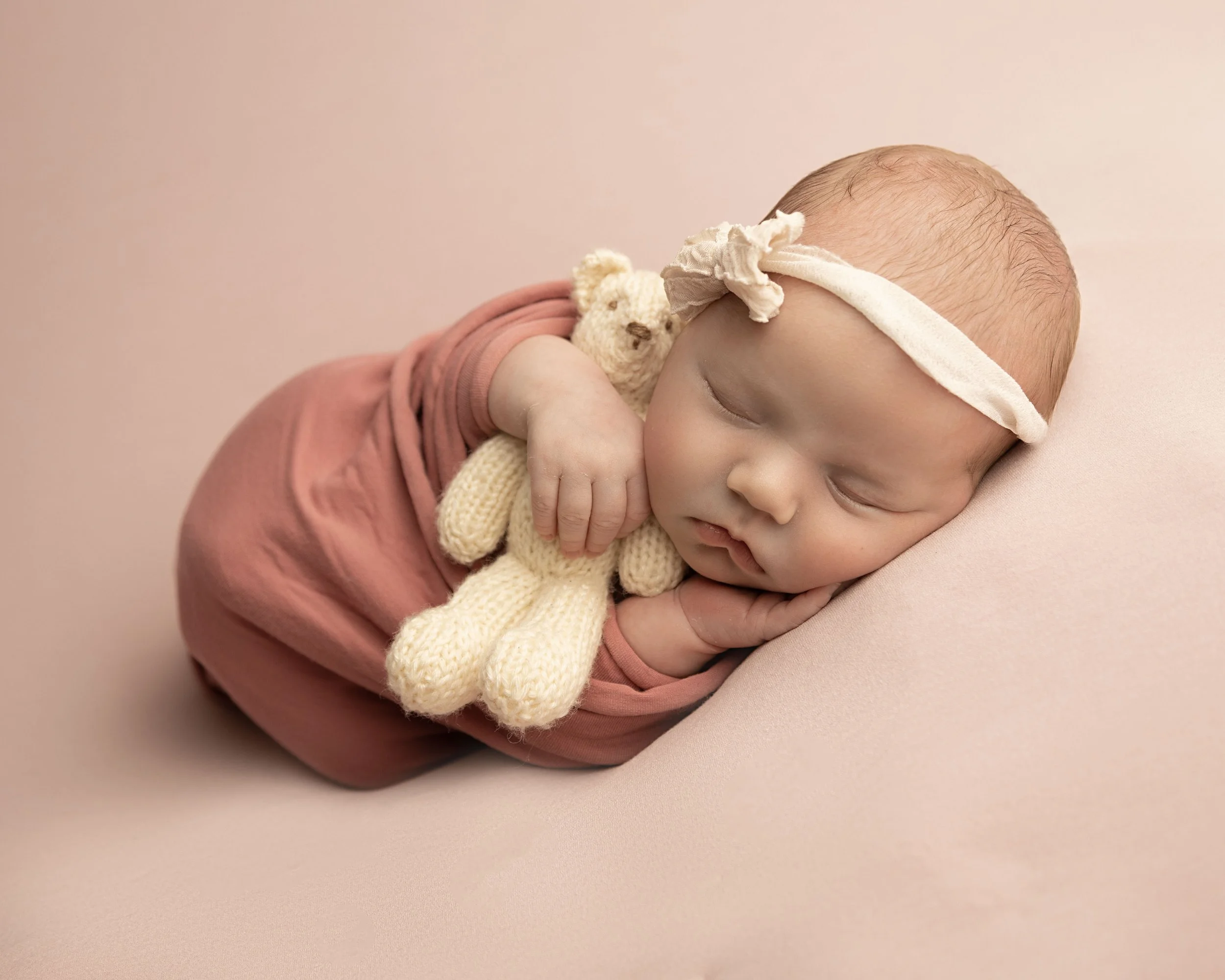 A sleeping baby with a headband, cuddling a knitted teddy bear on a soft pink background.