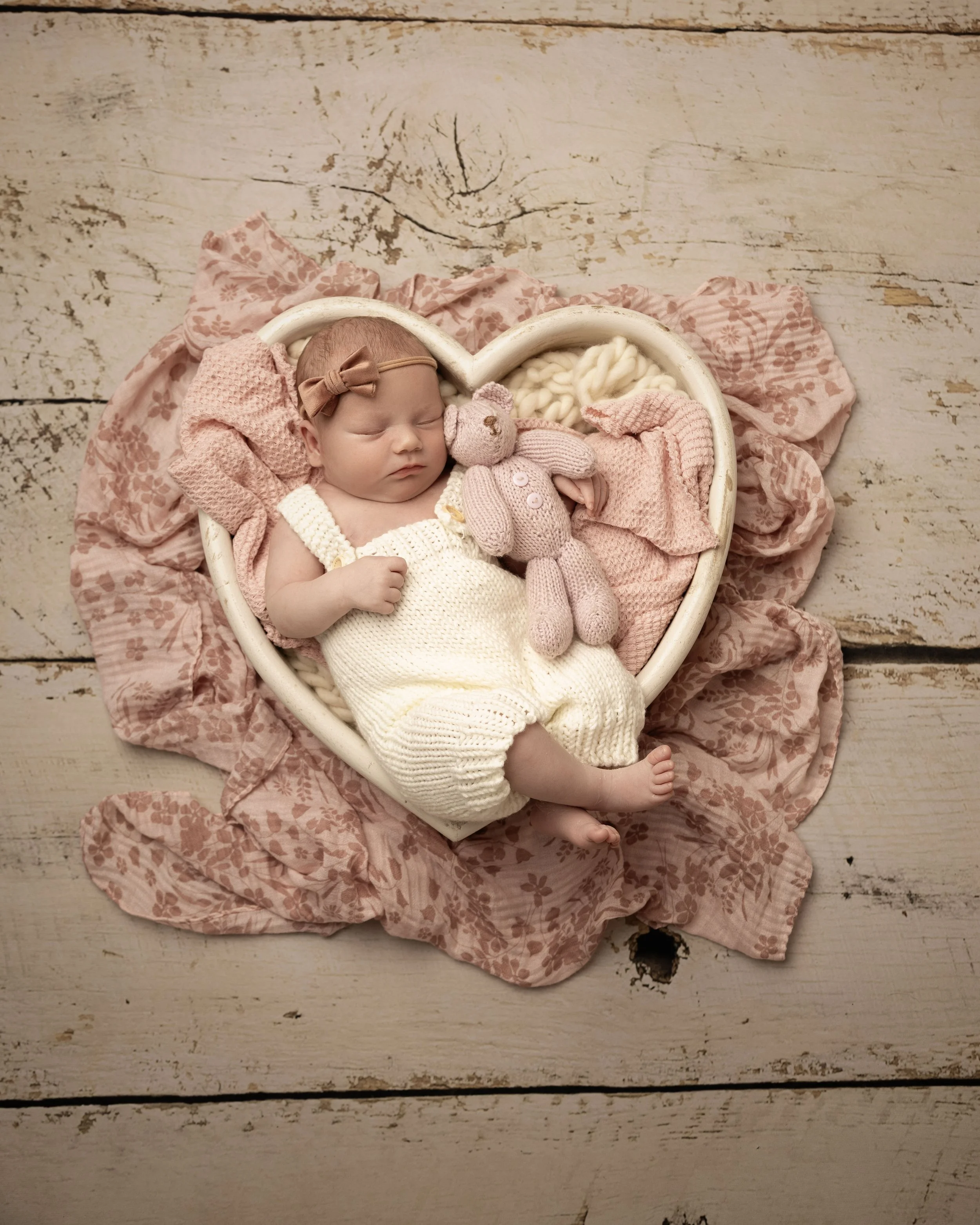 A sleeping baby girl lies in a heart-shaped basket with pink blanket and cream-colored knit clothing, holding a pink stuffed bunny, surrounded by a pink floral cloth on a white wooden floor.