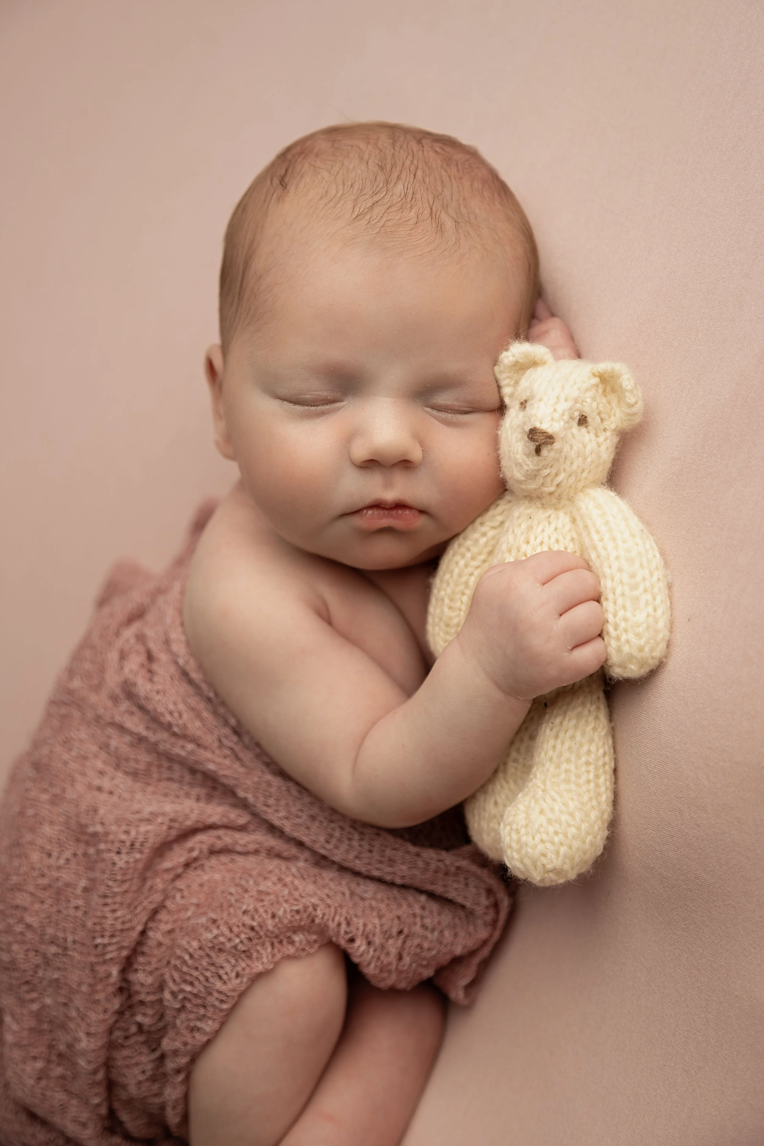 A sleeping baby cuddling a small knitted teddy bear, wrapped in a pink towel.