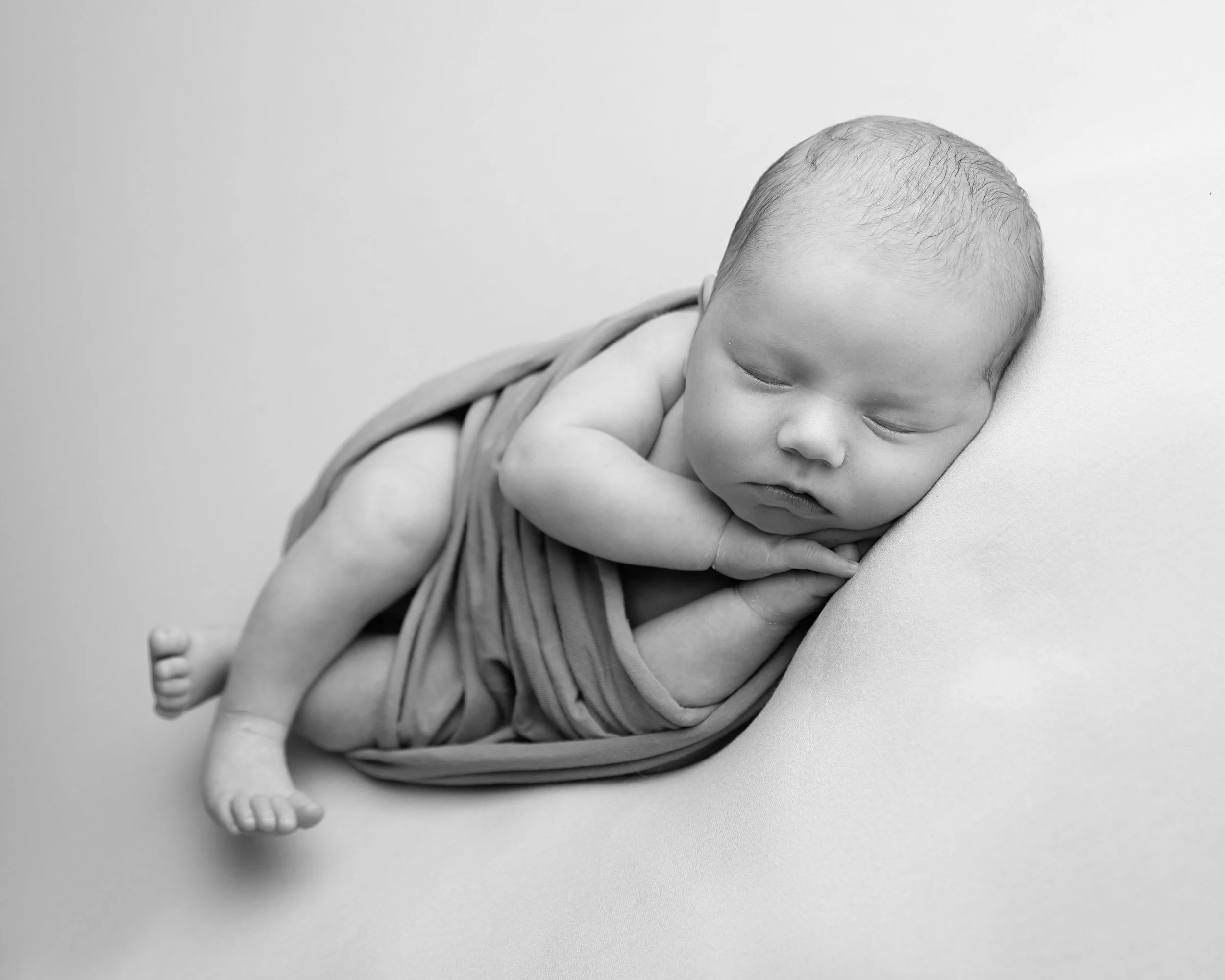 Black and white photo of a sleeping newborn baby swaddled in a blanket, lying on a soft surface.