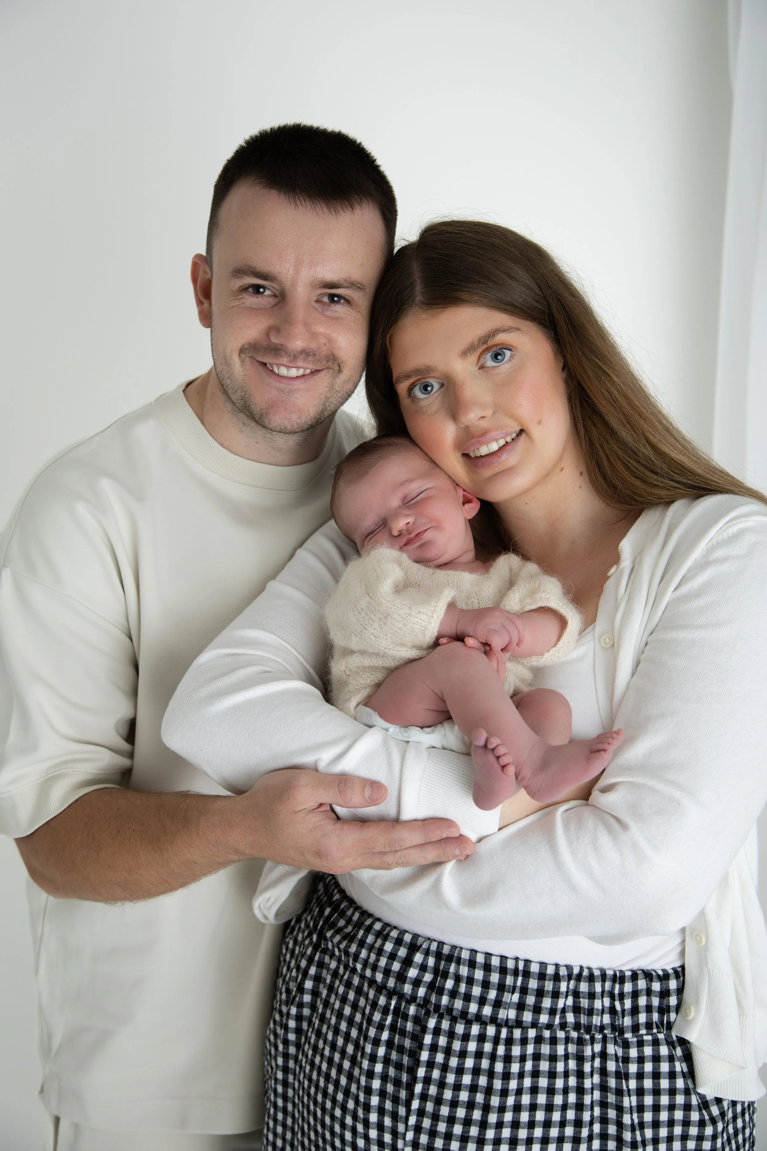 A happy family of three with a newborn baby, the father, mother, and baby all smiling and looking at the camera in a bright room.