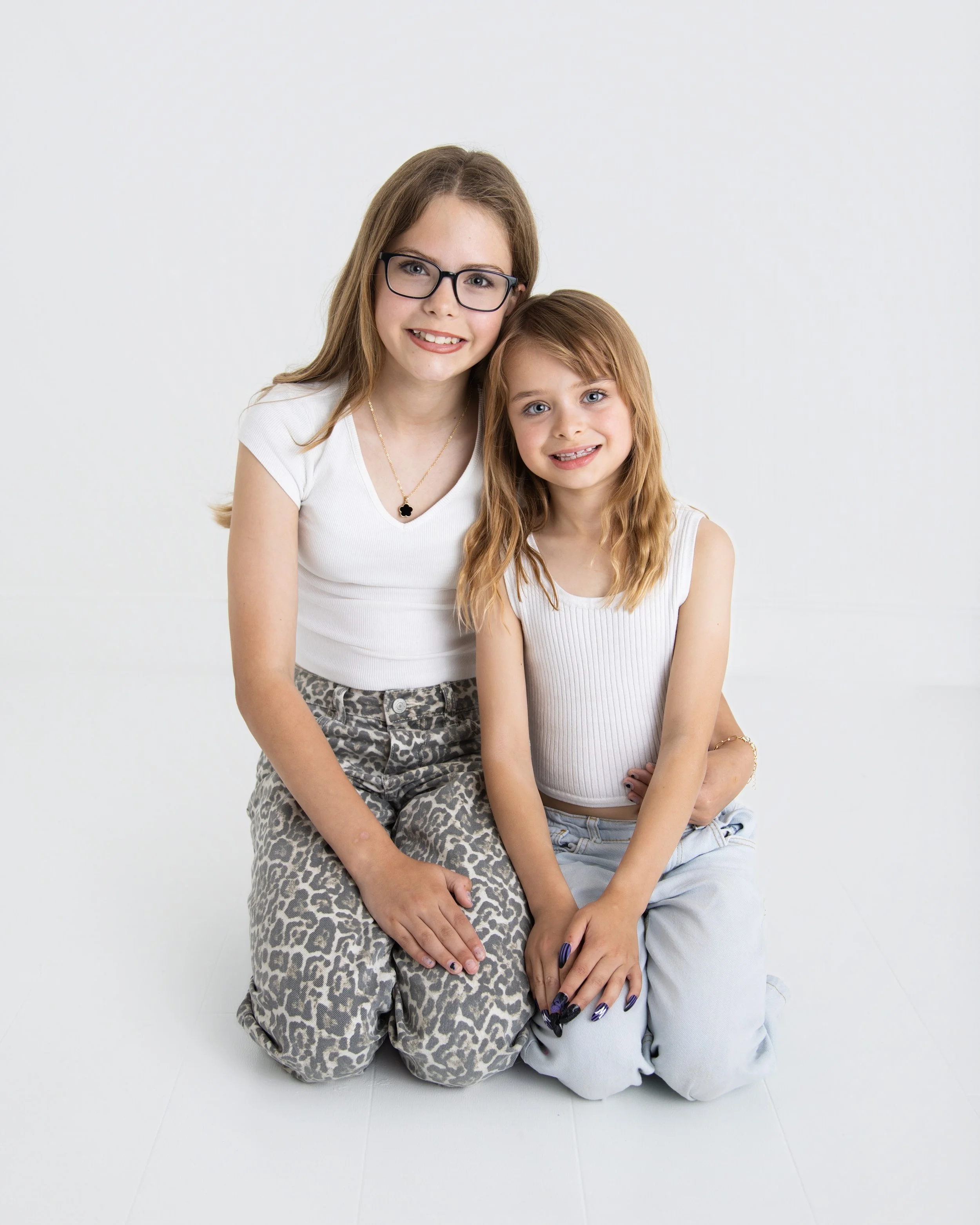 Two smiling girls, one older with glasses and one younger, sitting on the floor in front of a plain white background.