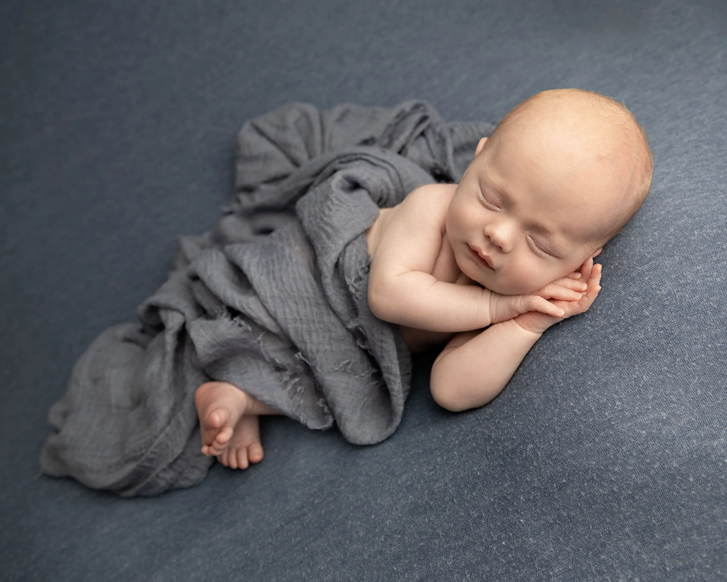A sleeping baby lying on a blue surface, partially wrapped in a gray blanket, with hands under their cheek and one leg slightly bent.