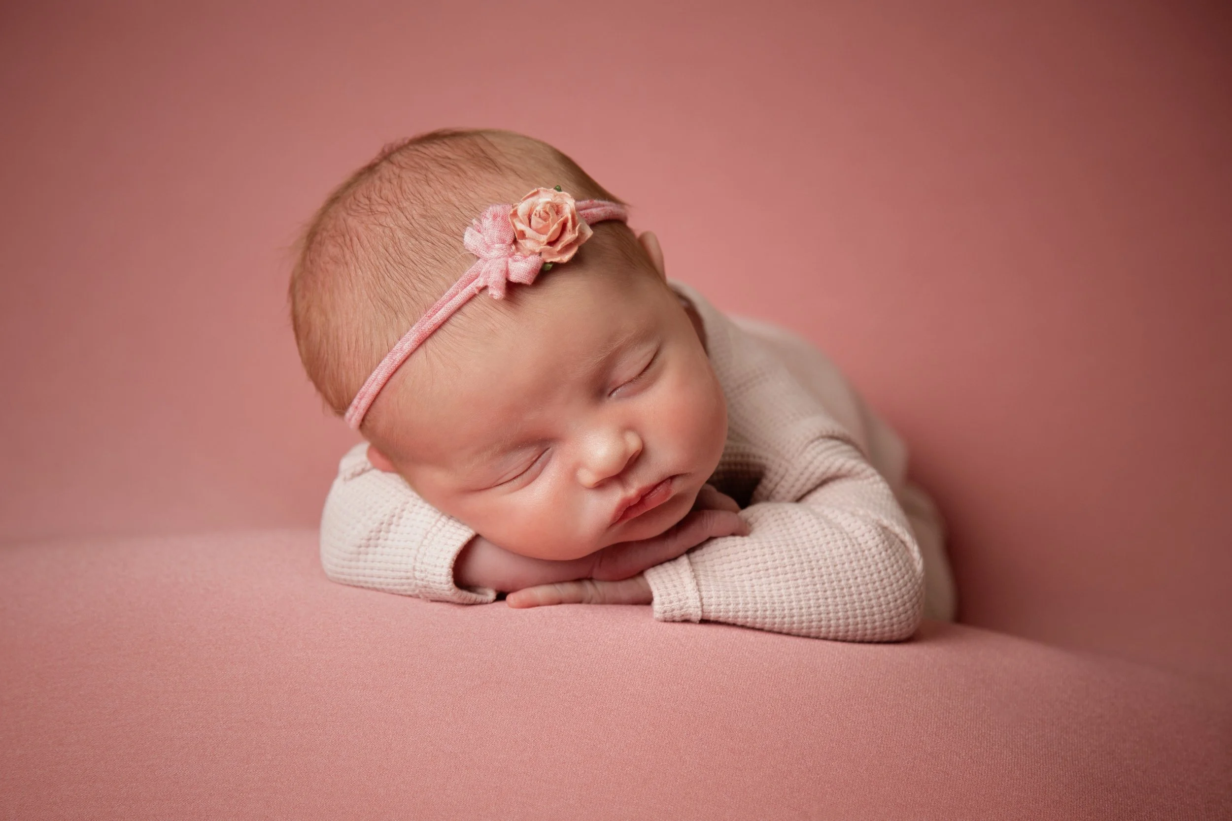 A sleeping newborn baby girl with a pink flower headband, resting her head on her arms on a pink surface.