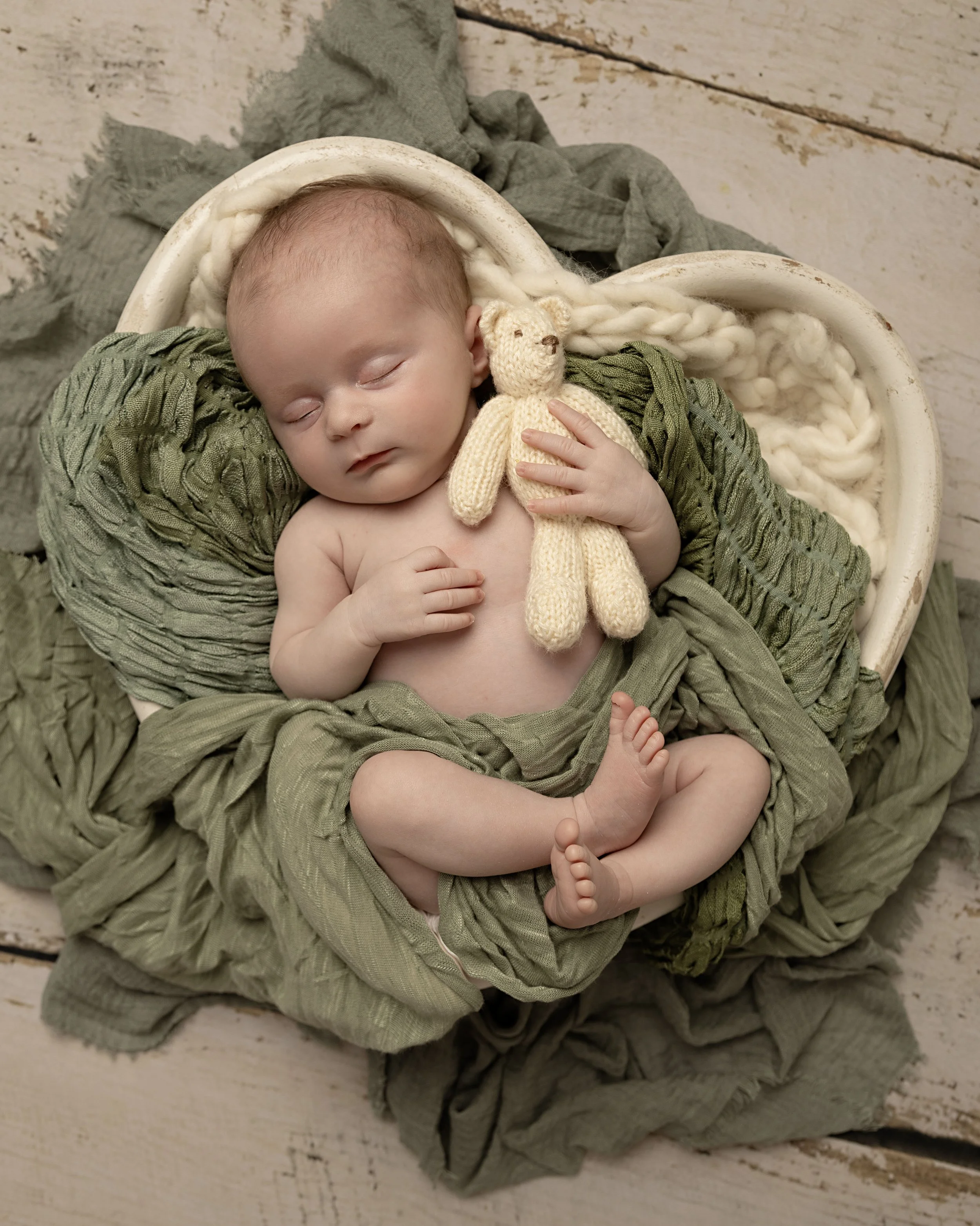 A sleeping baby with a knitted teddy bear, surrounded by green and beige fabrics on a wooden background.