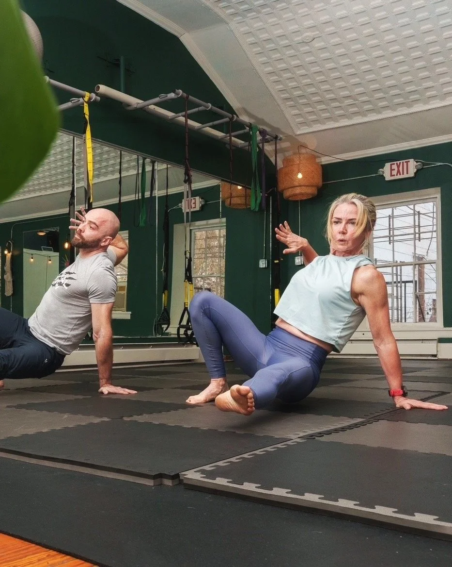 Two people exercising indoors on mats, performing mobility or yoga exercises, with straps hanging from the ceiling and green walls in the background.