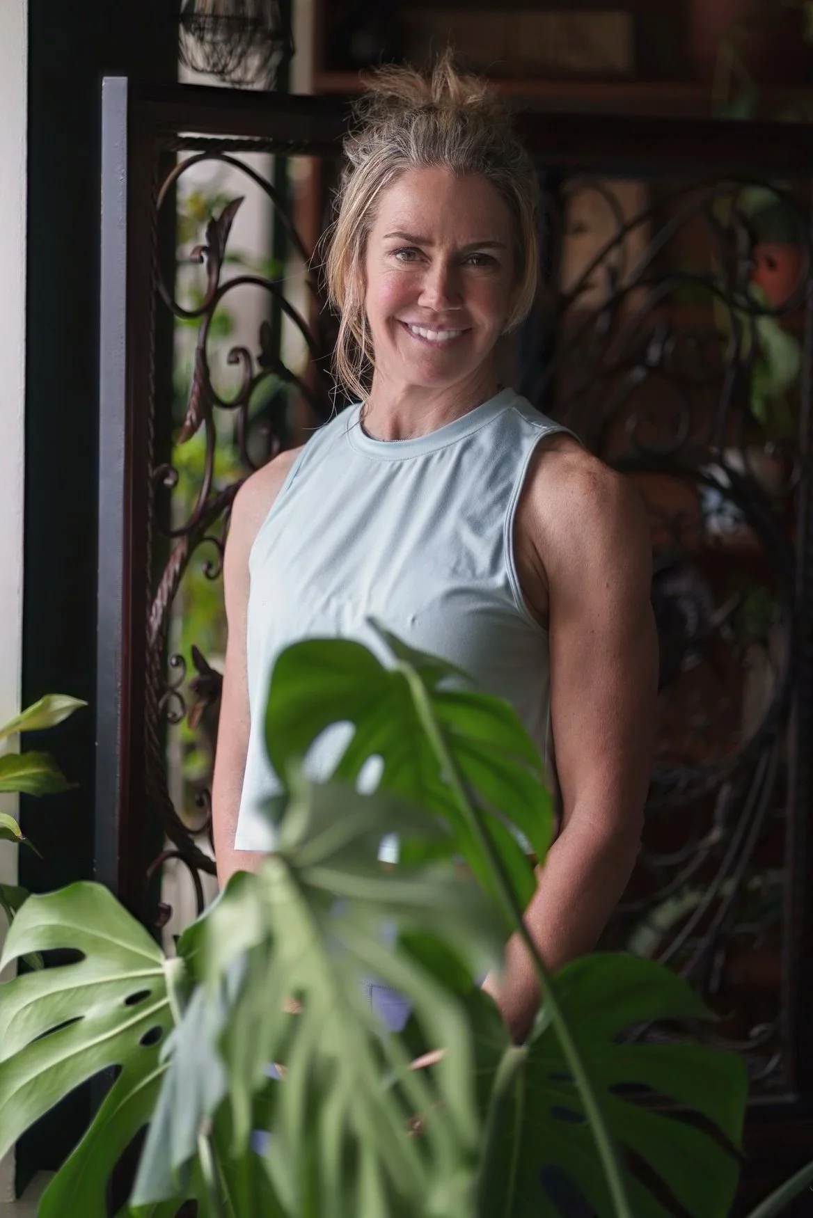 Person smiling in front of a decorative background and plants, wearing a sleeveless top.