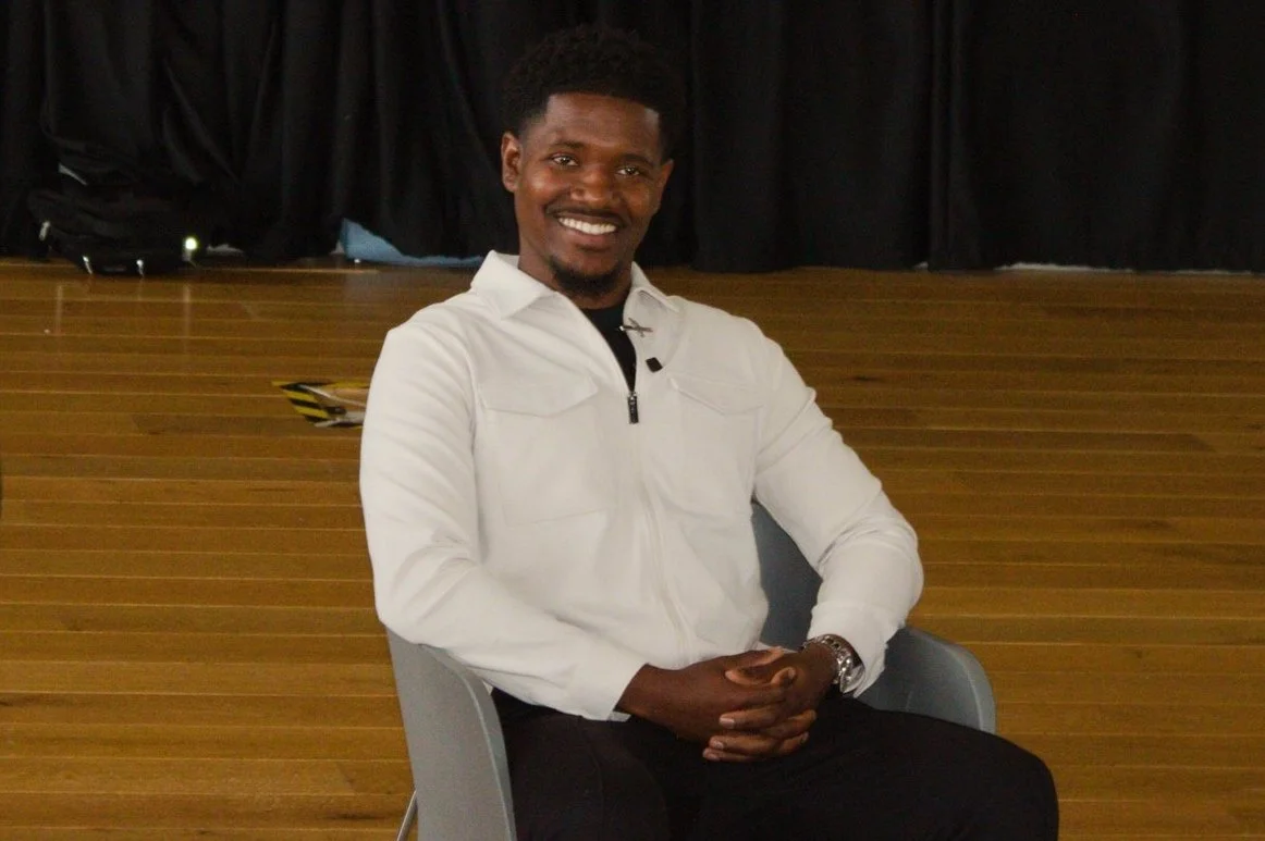 Smiling person in a white shirt sitting on a chair with a wooden floor and dark curtain backdrop.