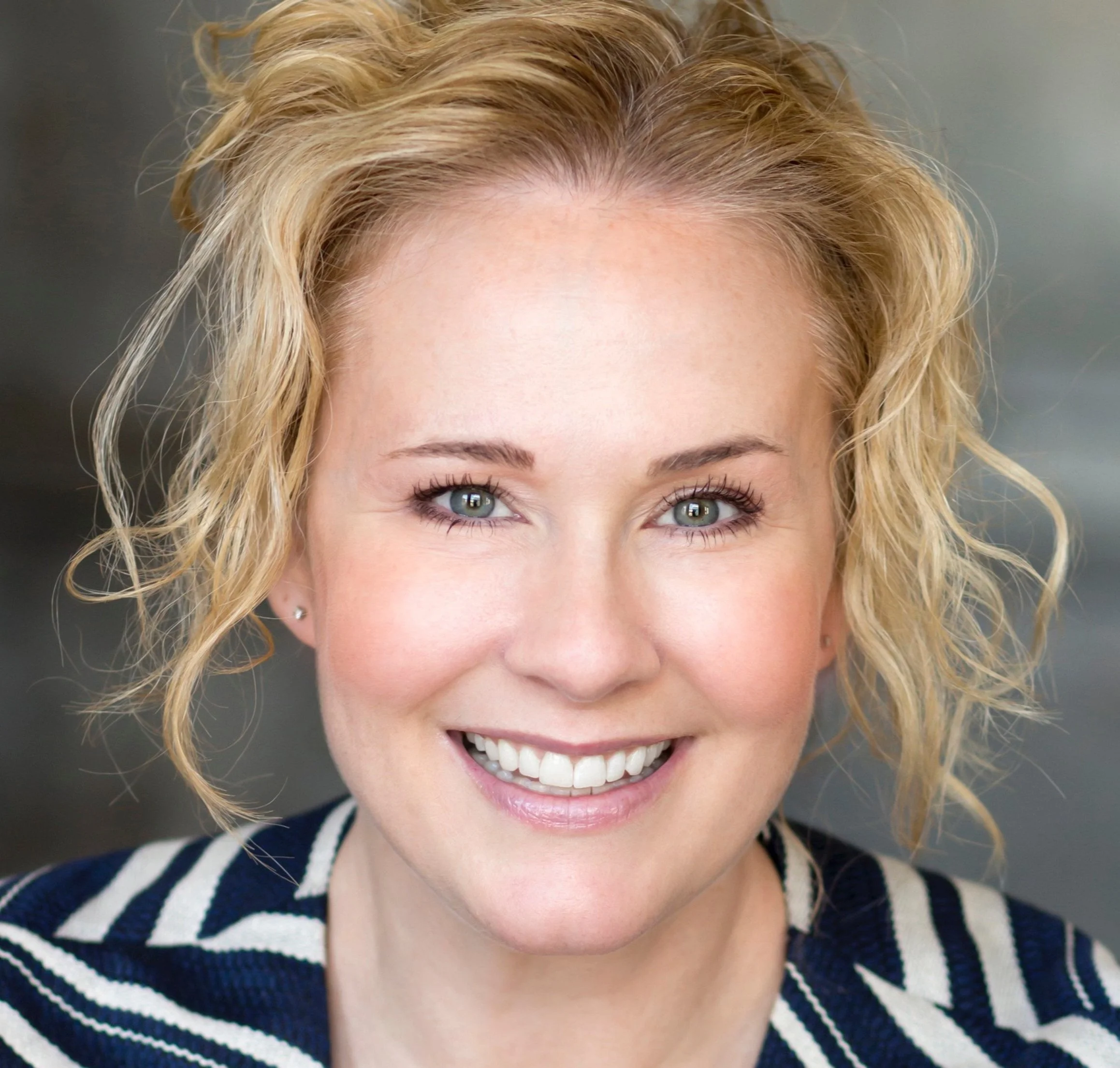Close-up of a smiling woman with blonde curly hair, blue eyes, and light makeup, wearing a navy blue and white striped top.