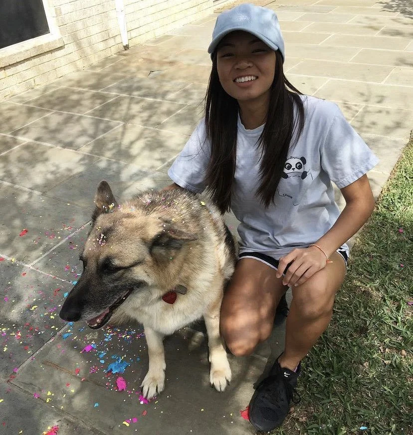 Woman kneeling beside a German Shepherd dog covered in colorful confetti. She is wearing a light blue baseball cap, a t-shirt with a panda, and has a smile. The setting is outdoors on a paved area with a grassy edge.