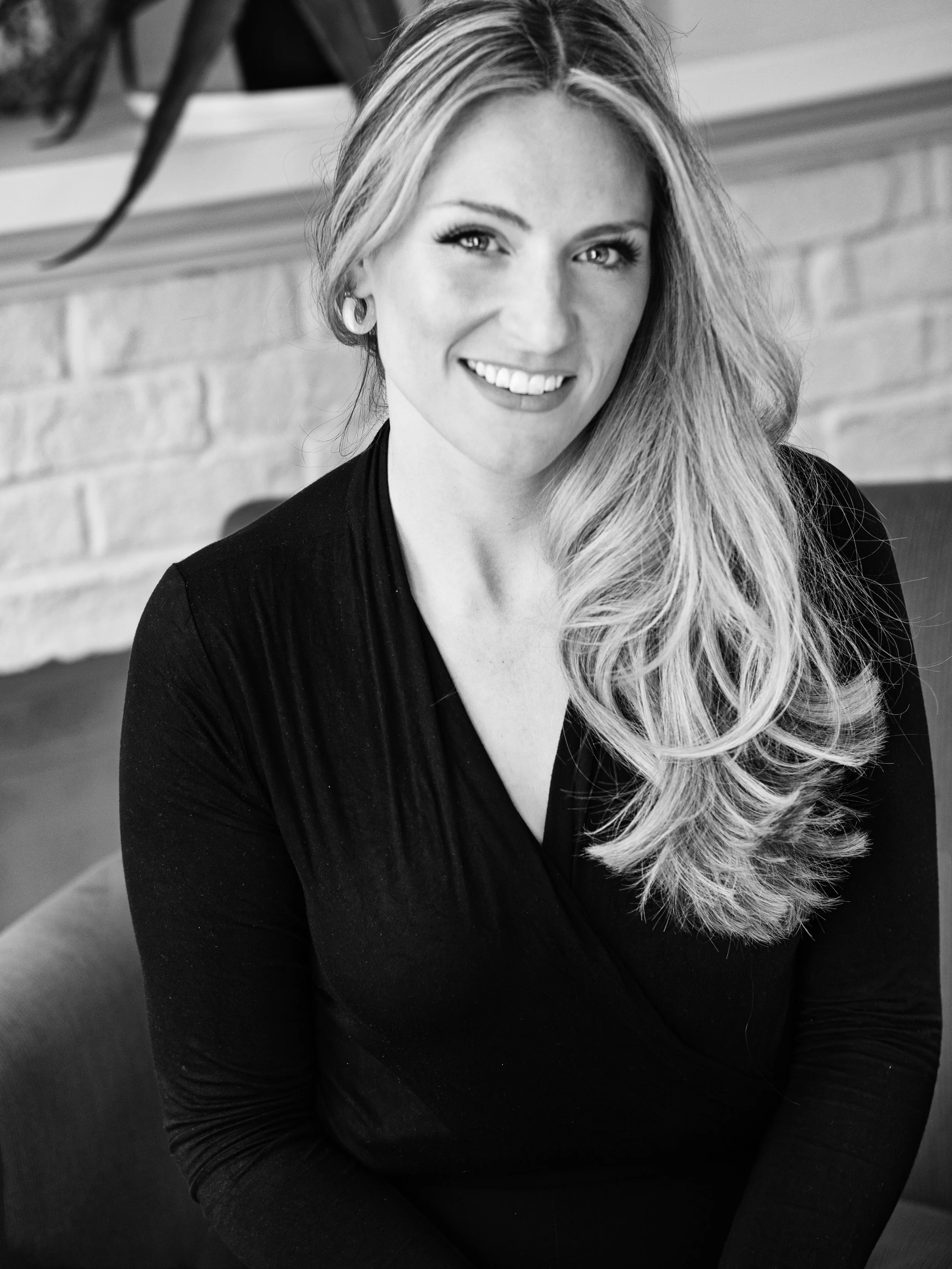 Black and white portrait of a smiling woman with long hair wearing a dark top, sitting indoors with a brick wall background.