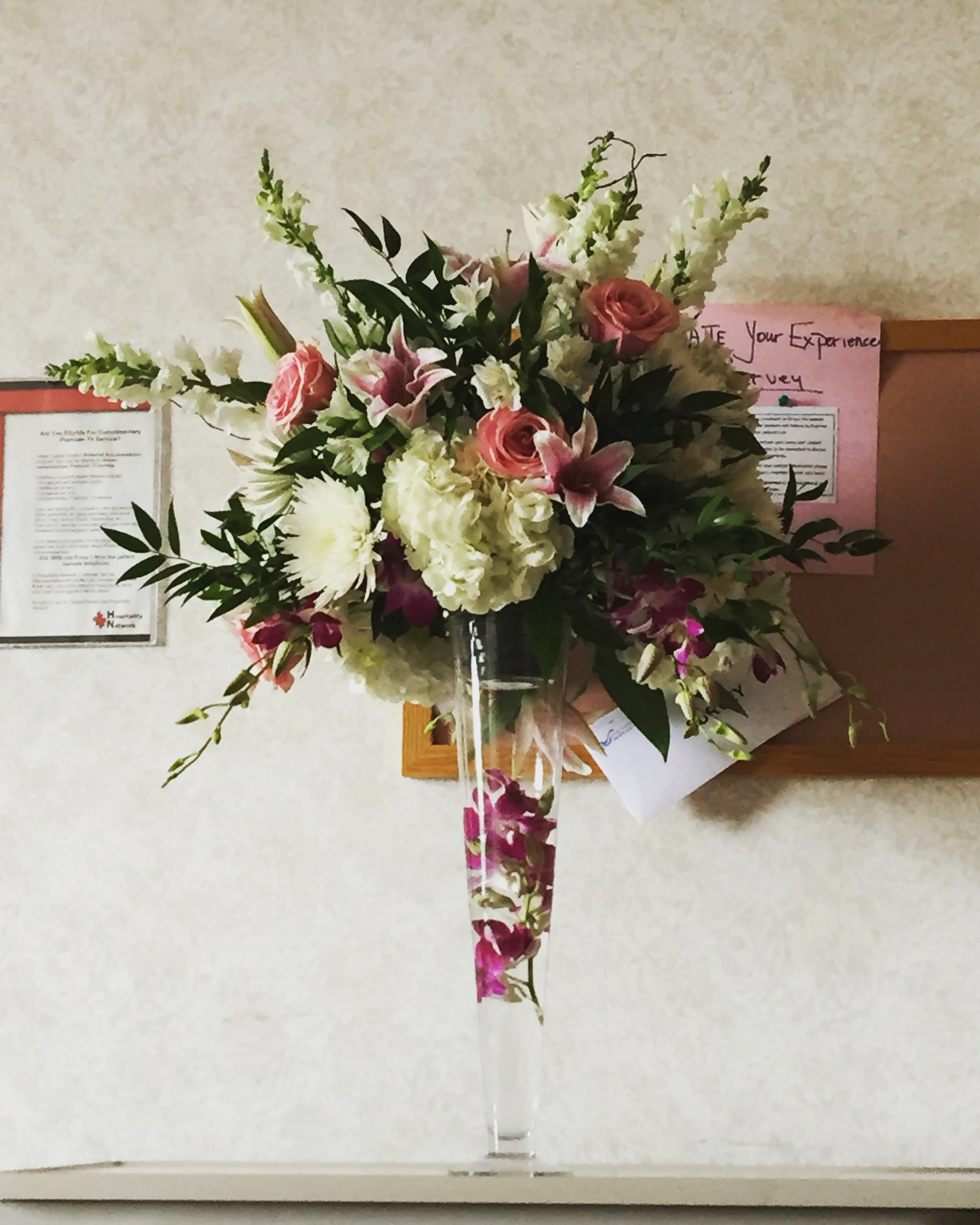 Elegant floral arrangement with pink roses, white lilies, and assorted greenery in a tall glass vase on a table, surrounded by documents on a notice board in the background.