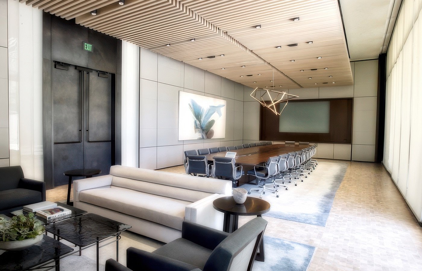 Modern conference room with a long table surrounded by gray office chairs, a large wall-mounted screen, artistic lighting fixture, and comfortable sitting area with sofas and armchairs.