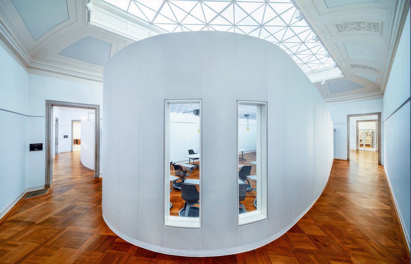 Modern interior space with a circular white partition wall with three vertical windows, surrounding a small conference room with chairs and tables. Wood flooring and a skylight ceiling with intricate molding details.