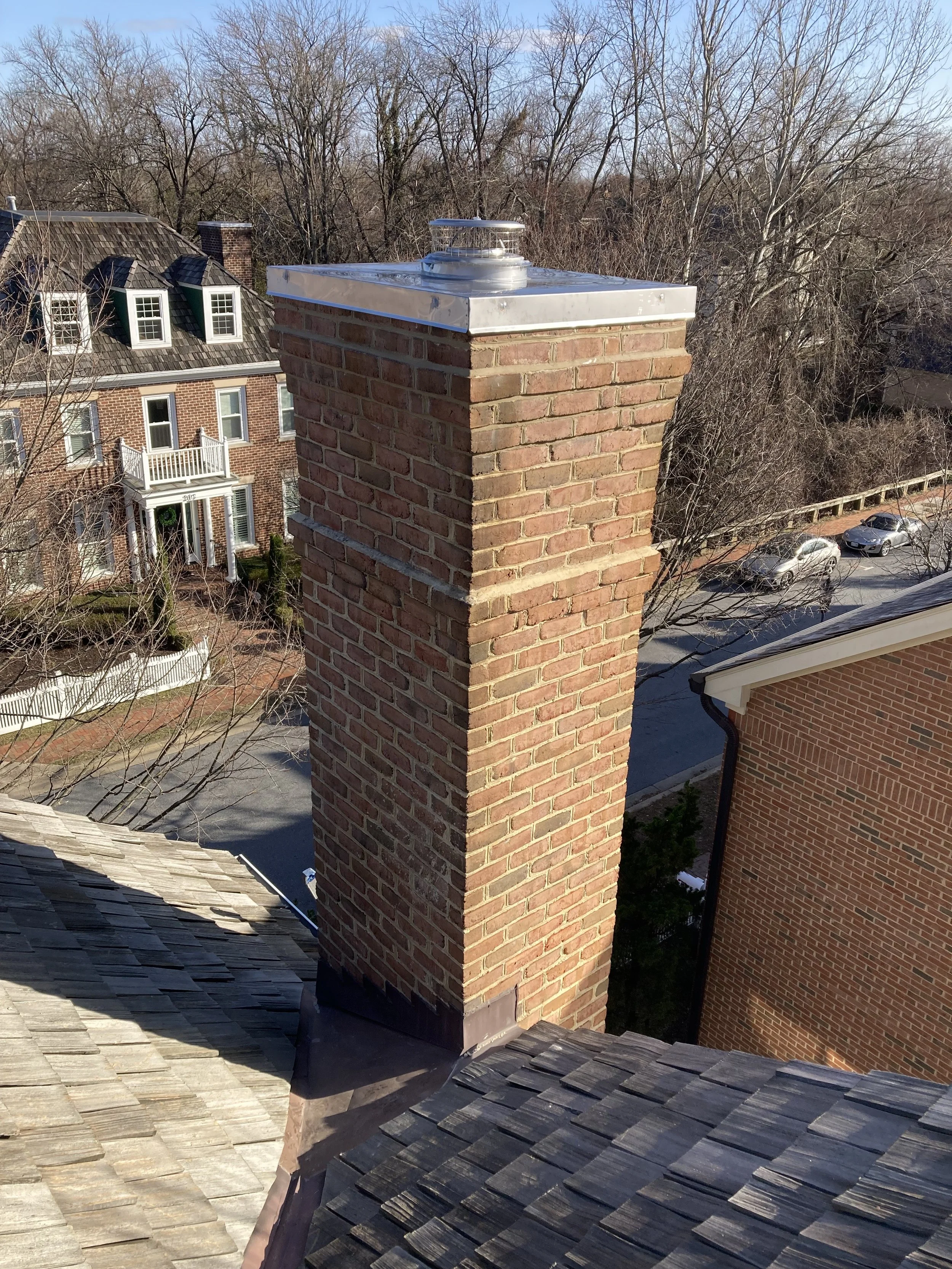 Brick chimney on a shingled roof in a suburban neighborhood with houses and bare trees in the background.