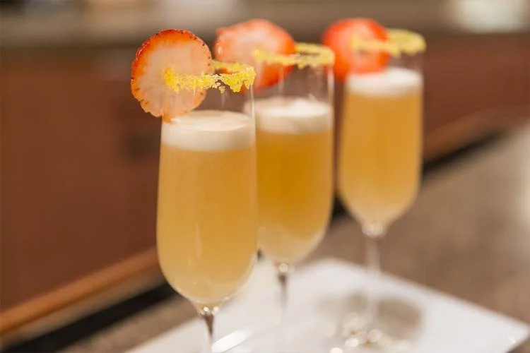 Three champagne glasses filled with a bubbly yellow drink, garnished with a slice of strawberry and a twist of lemon peel.