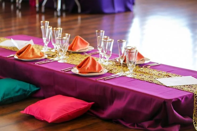 A banquet table with purple tablecloth, gold table runner, colorful napkins, glassware, place settings, and floor cushions on a wooden floor.