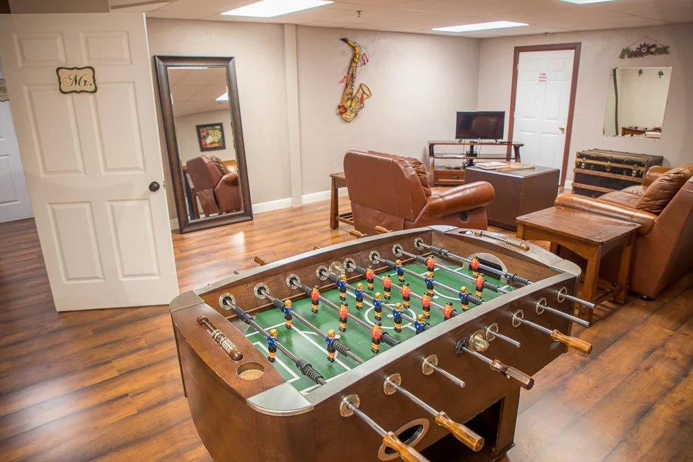 An indoor game room with a foosball table in the foreground, leather armchairs and a coffee table, a flat-screen TV on a stand, a large mirror, a wall decoration, and a door in the background.
