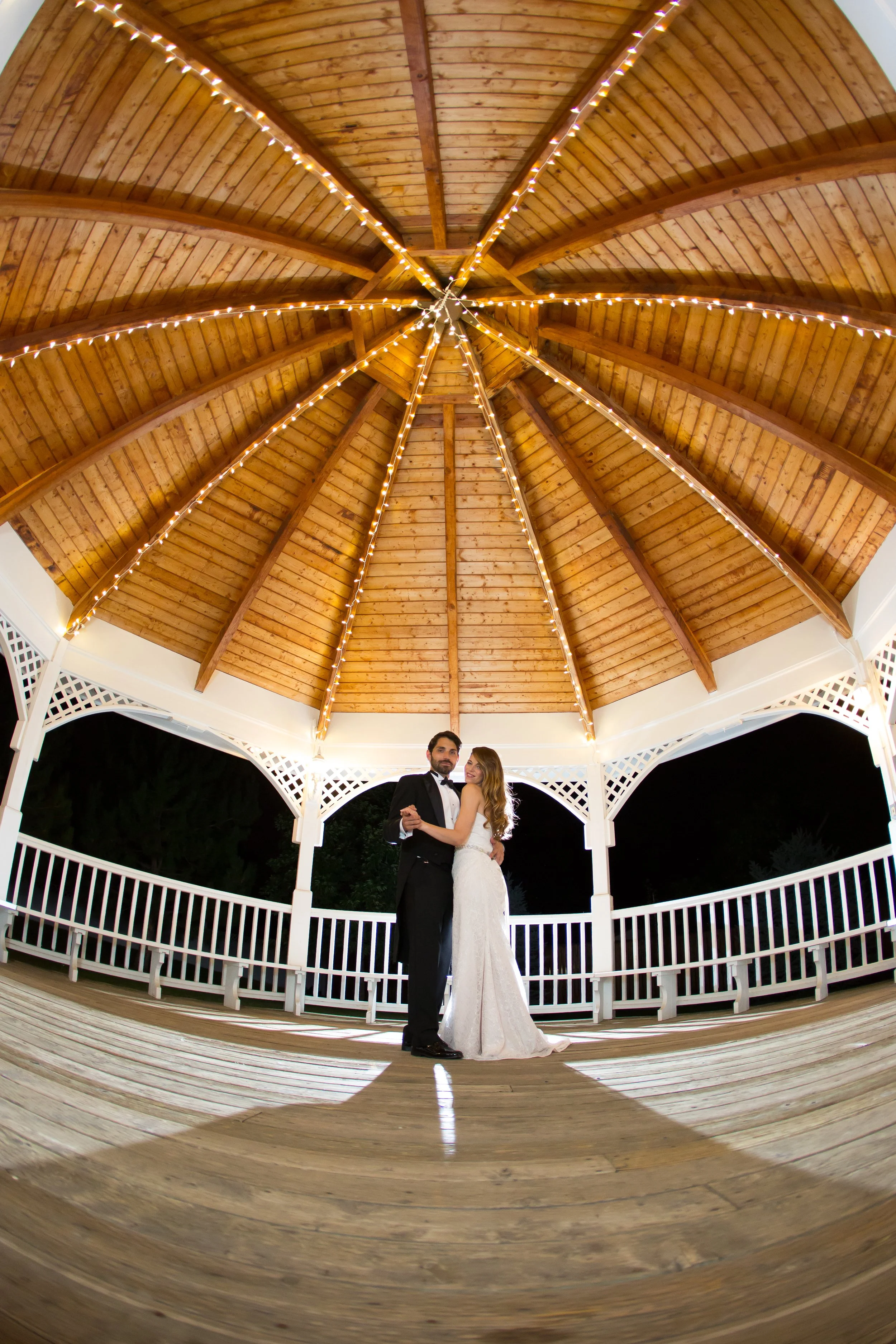 A bride and groom dancing in an outdoor gazebo at night, with a wooden ceiling and string lights overhead.