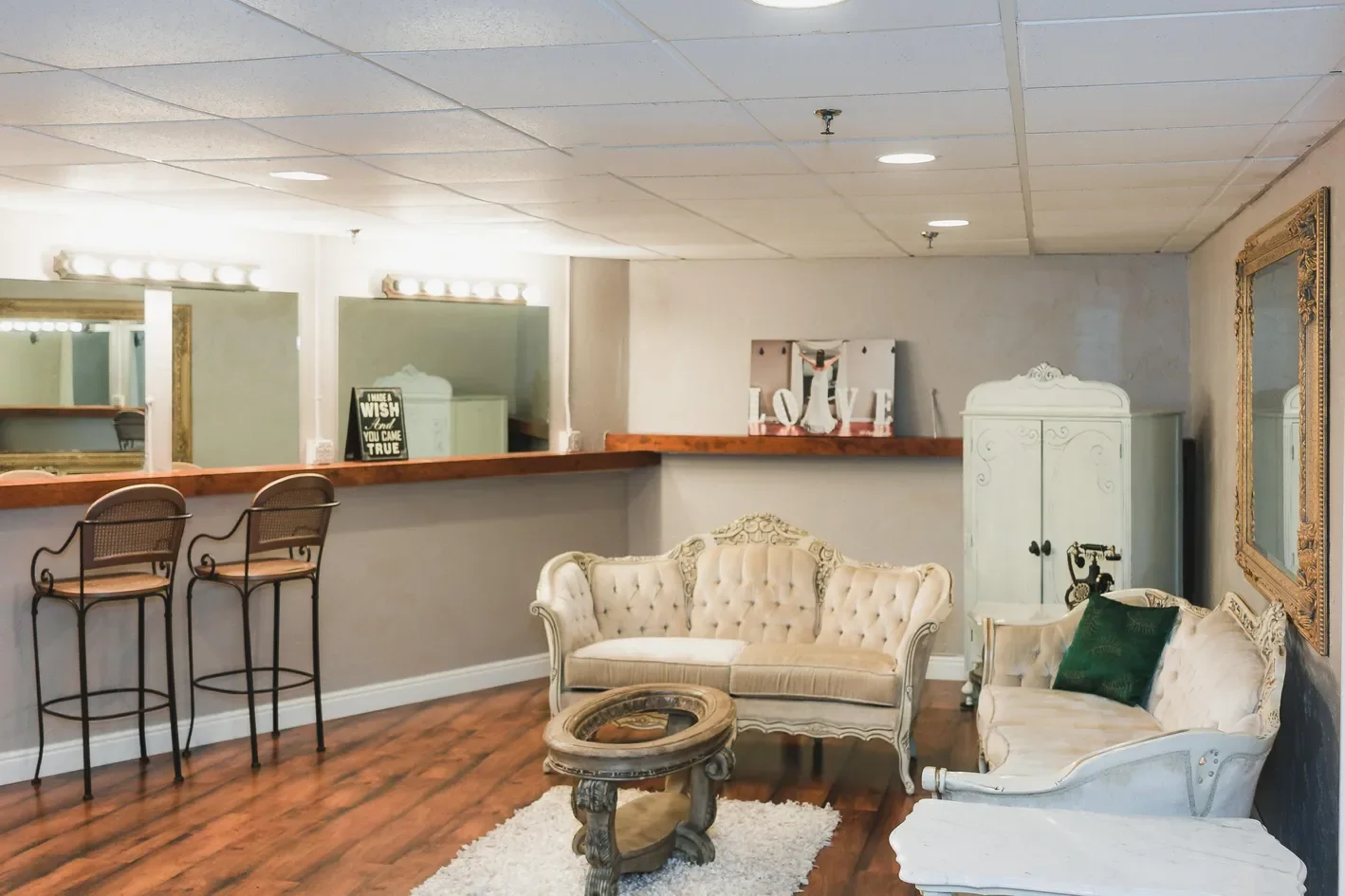 Living room with vintage-style furniture, including a tufted cream sofa, a matching armchair, an ornate coffee table, a white cabinet, and a large framed mirror on the wall. There is a bar area with two chairs in the background and wall-mounted mirro