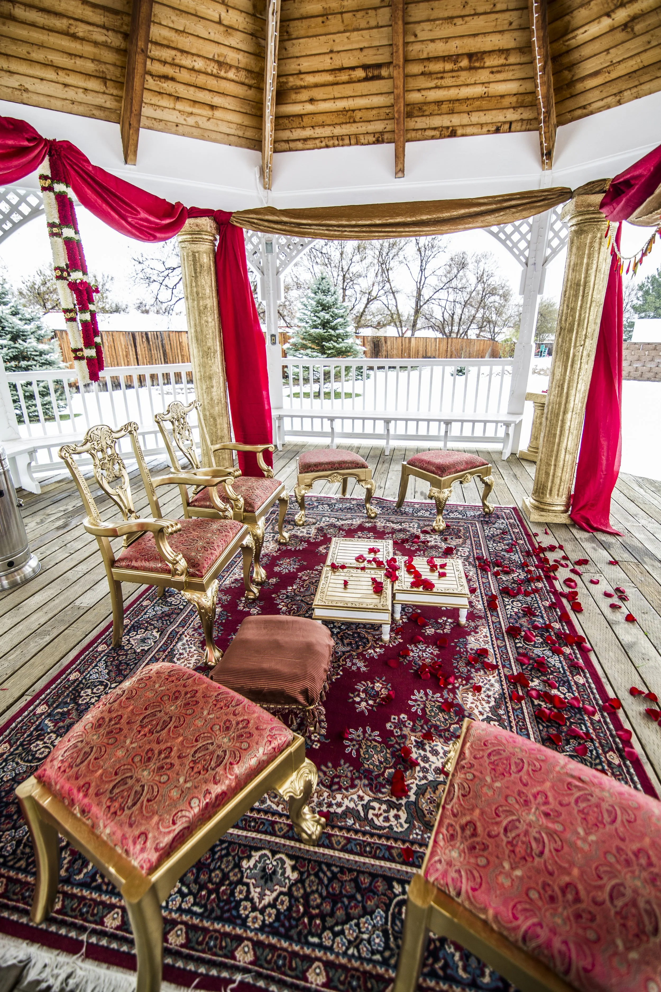 A decorated outdoor gazebo with ornate chairs, a patterned rug, and rose petals scattered on a small table, with snow outside and red drapes hanging from the columns.