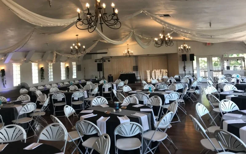 Wedding reception hall decorated with draped white fabric on the ceiling, chandeliers, and string lights. Multiple round tables with black tablecloths and white chairs are arranged throughout the room. Large illuminated 'LOVE' sign on the stage at th