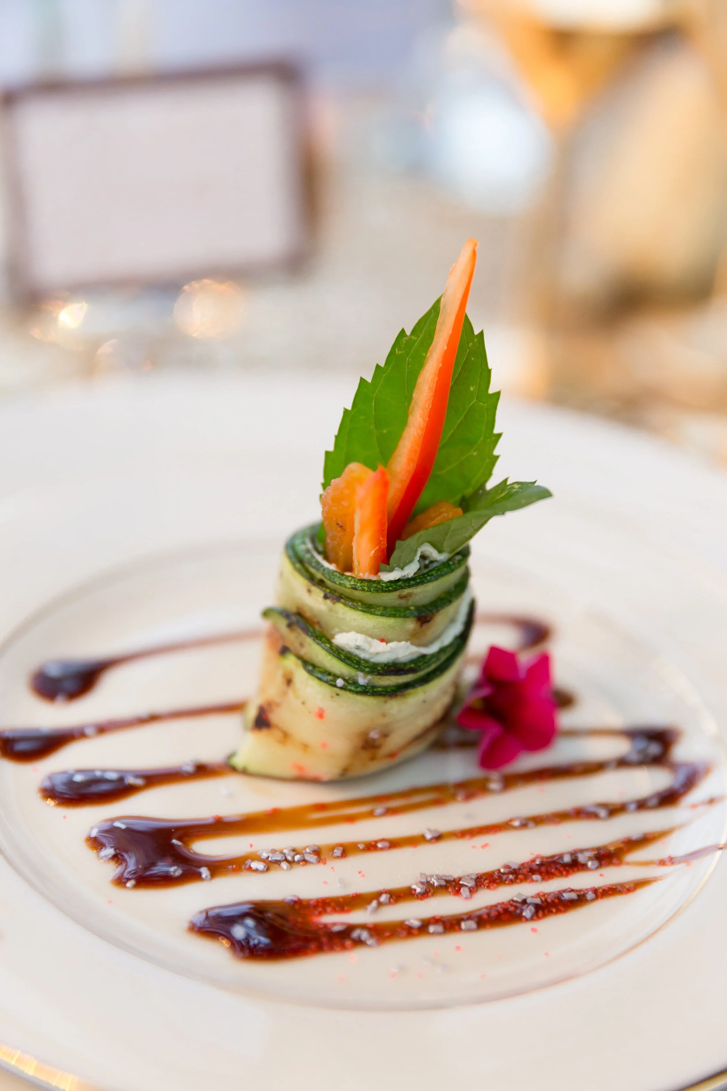 An elegantly plated vegetable dish featuring thinly sliced zucchini rolled and topped with a leaf of fresh green herbs, garnished with a pink flower, and decorated with drizzles of balsamic glaze and sauce on a white plate.