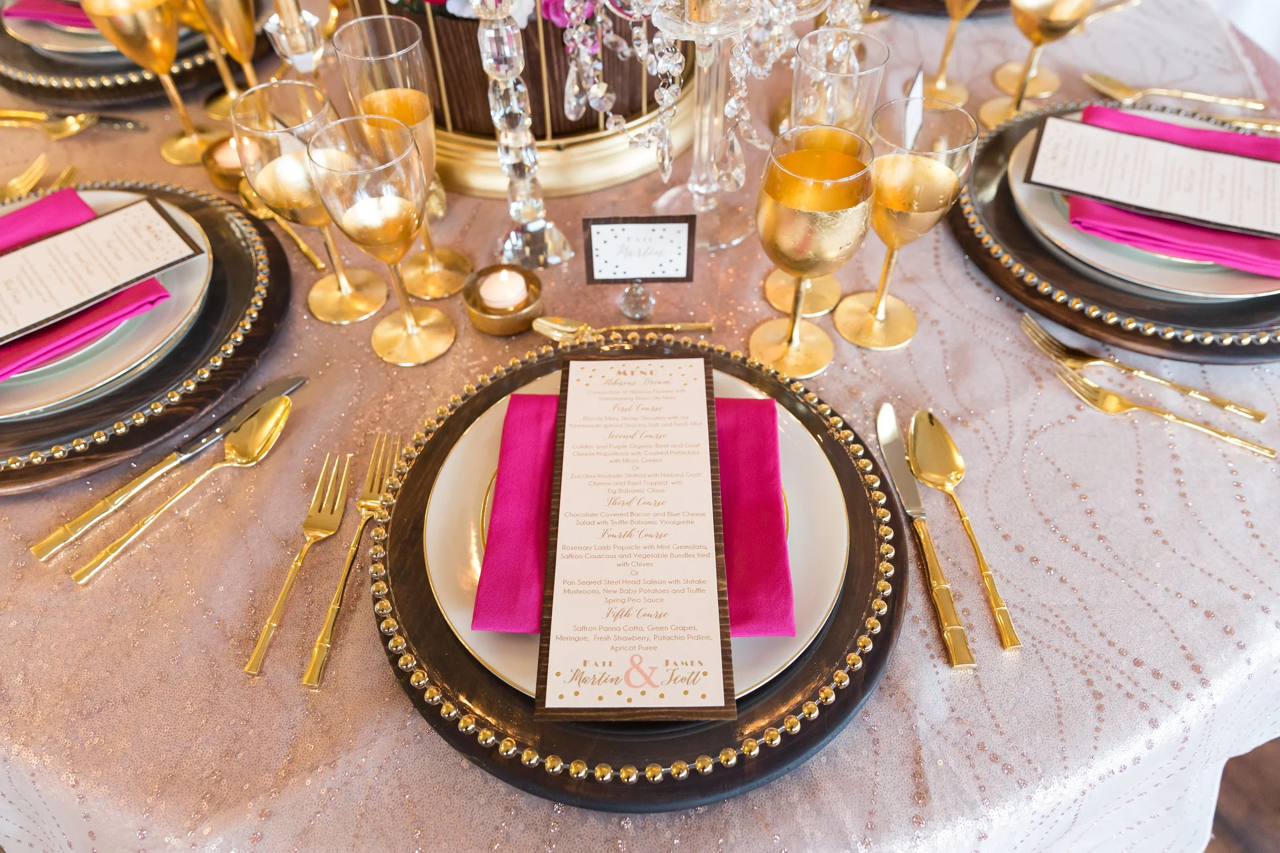 Elegant wedding or special event table setting with black and gold beaded chargers, pink napkins, gold flatware, champagne glasses with gold rims, a decorative chandelier centerpiece, and a detailed menu card on each plate.