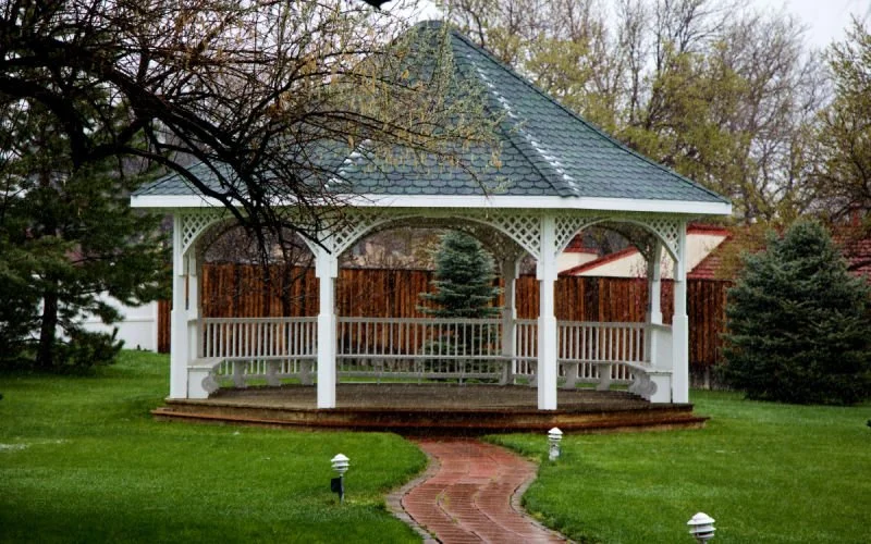 White gazebo with a dark gray roof on a grassy lawn, surrounded by trees and a brick pathway leading to it.