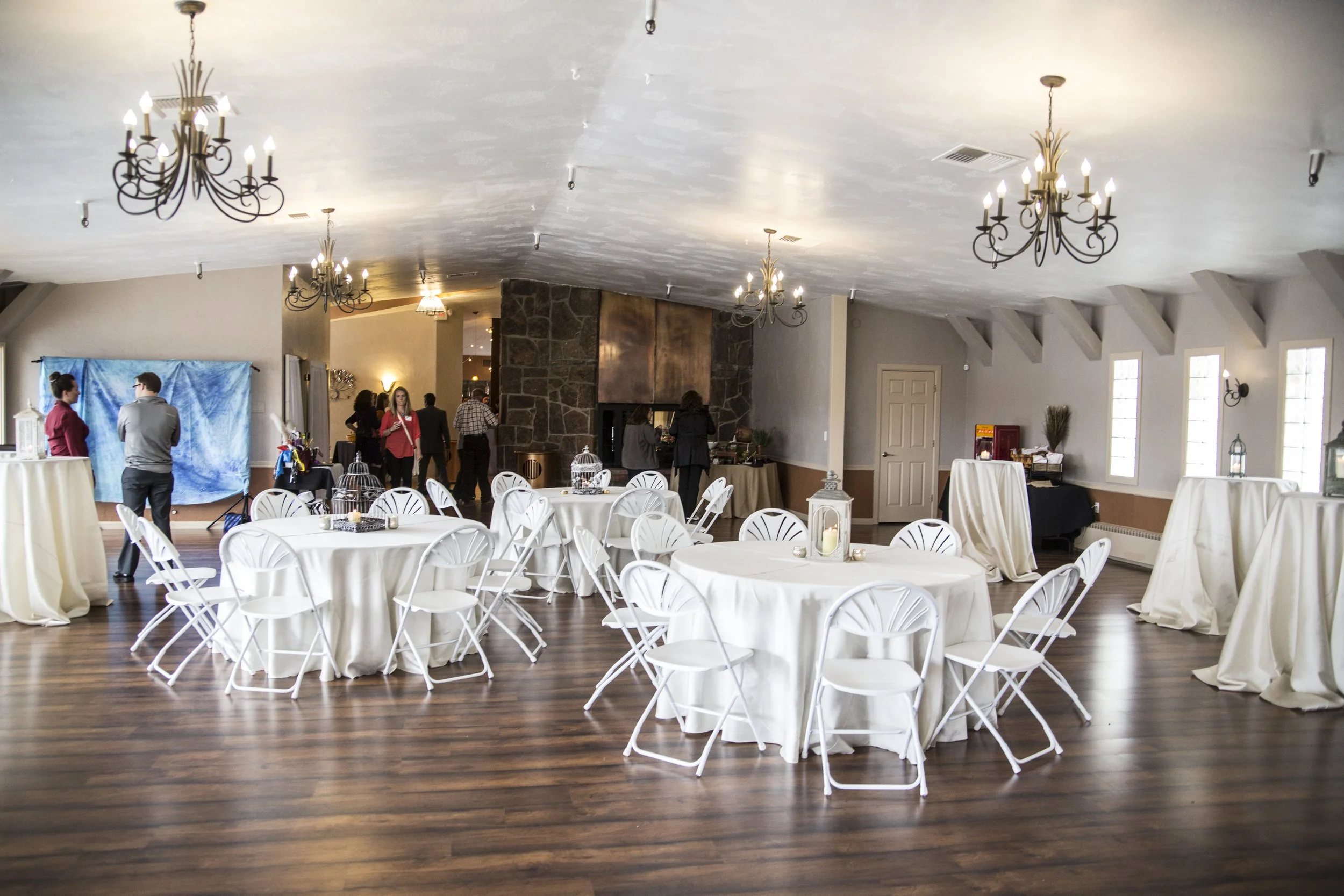 Event space decorated with round tables covered in white tablecloths, white chairs, chandeliers, and some people setting up or mingling. There are also high tables draped with cloth, a stone fireplace, and large windows letting in natural light.