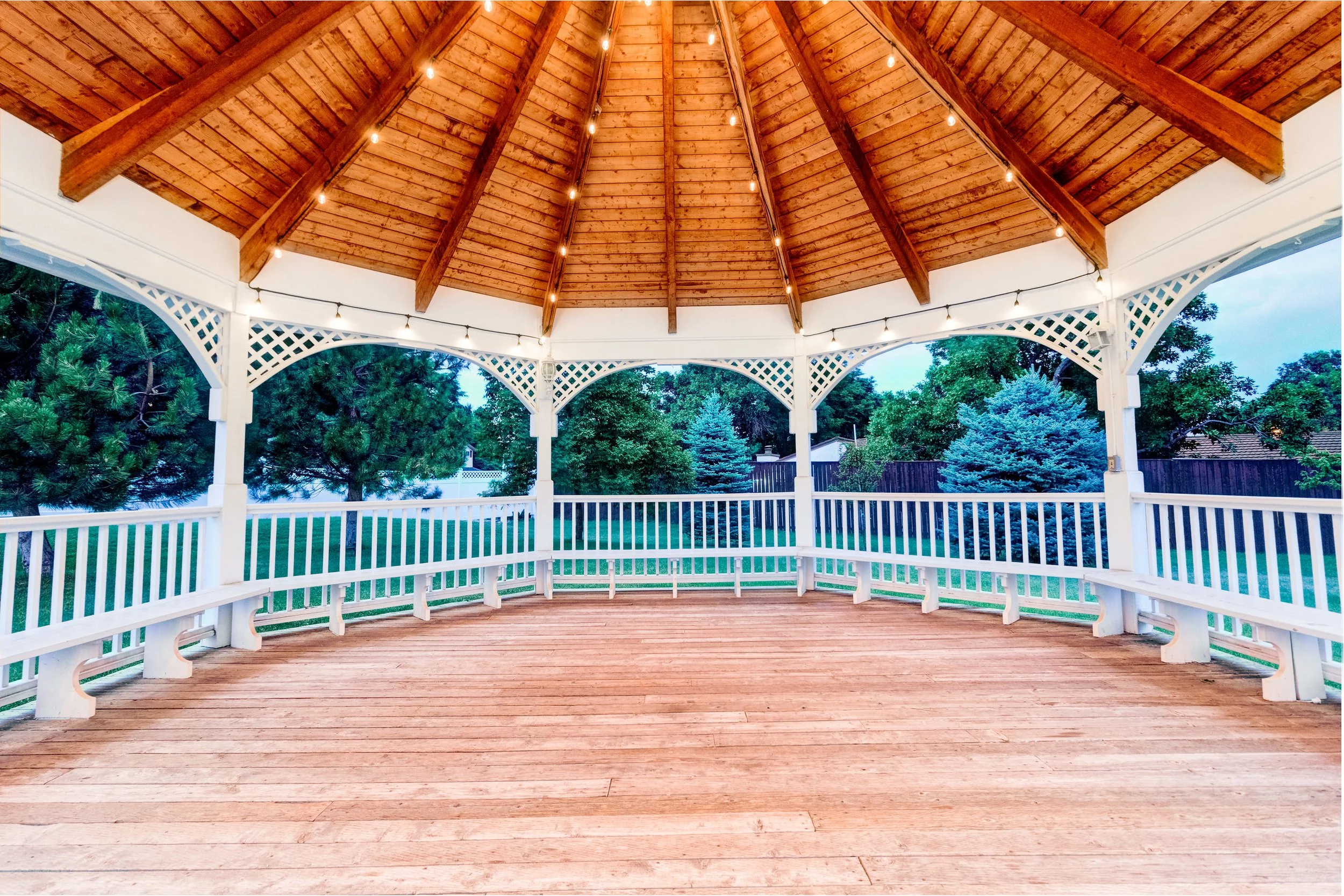 View from inside an outdoor gazebo with a wooden floor, white railings, a wooden ceiling with string lights, and trees and houses in the background.
