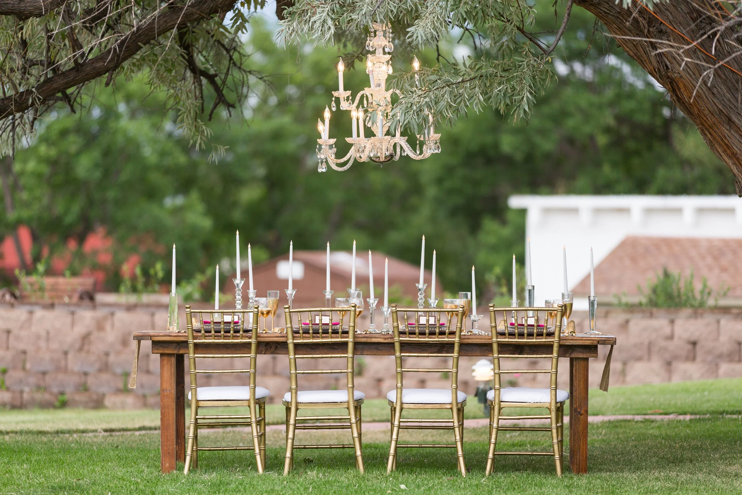 A decorated outdoor dining table with gold chairs, a chandelier hanging from tree branches, and lit candles, set in a garden with grass, trees, and buildings in the background.