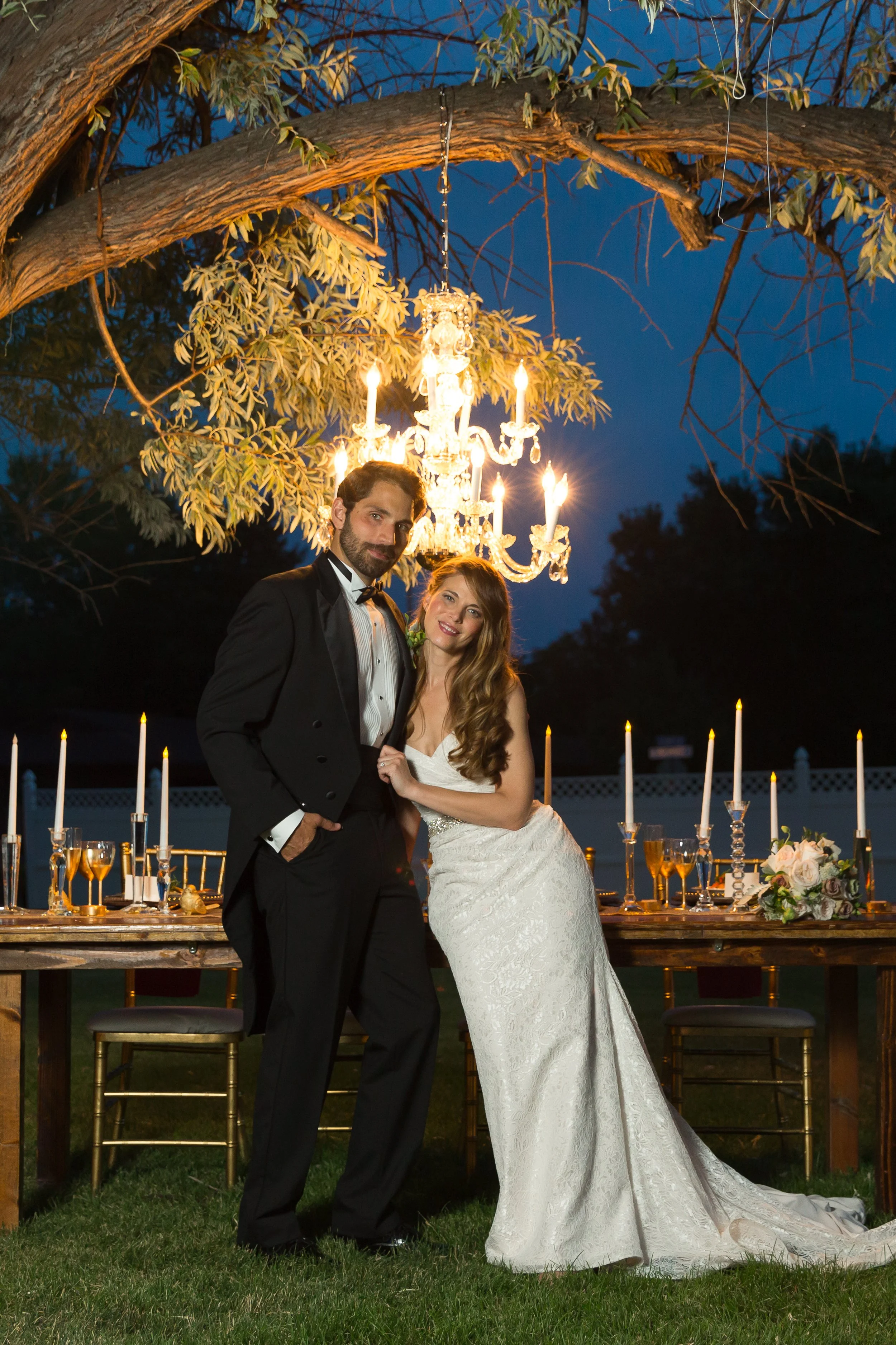 Bride and groom in formal attire at an outdoor evening wedding reception with a chandelier hanging from a tree and decorated table with candles and flowers.
