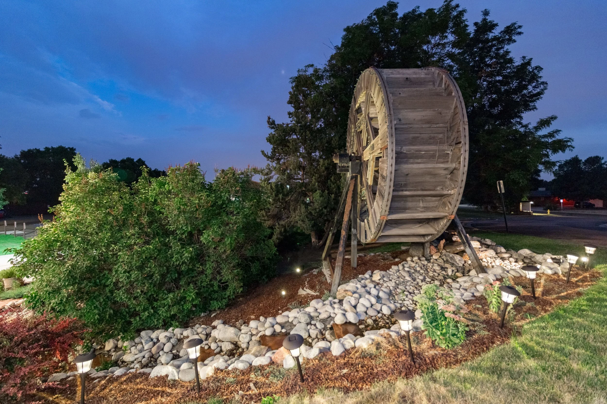 A vintage wooden water wheel mounted on supports in a landscaped garden at dusk, with garden lights illuminating rocks and plants.