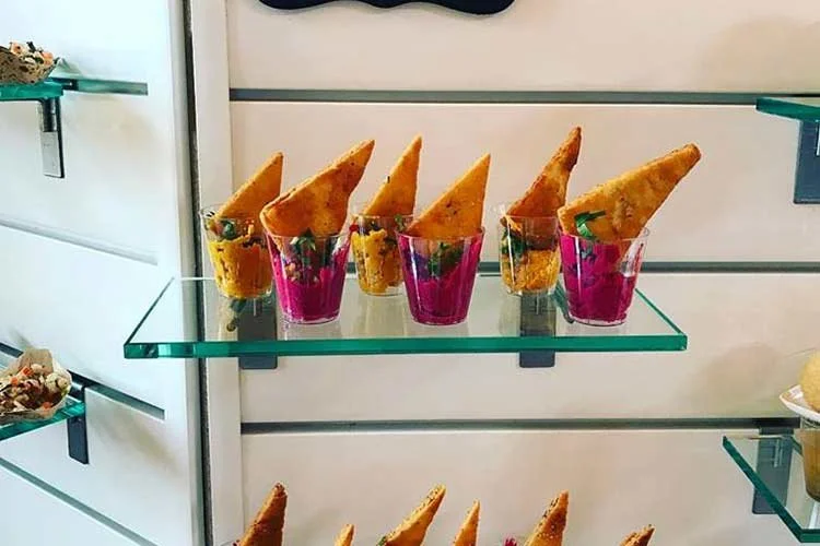 Six individual servings of fried appetizer wraps with green and purple ingredients, placed in pink transparent cups on a glass shelf.