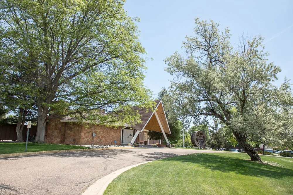 A building with a brown stone exterior and a steep triangular roof surrounded by green trees and a well-maintained lawn in a park-like setting on a clear day.
