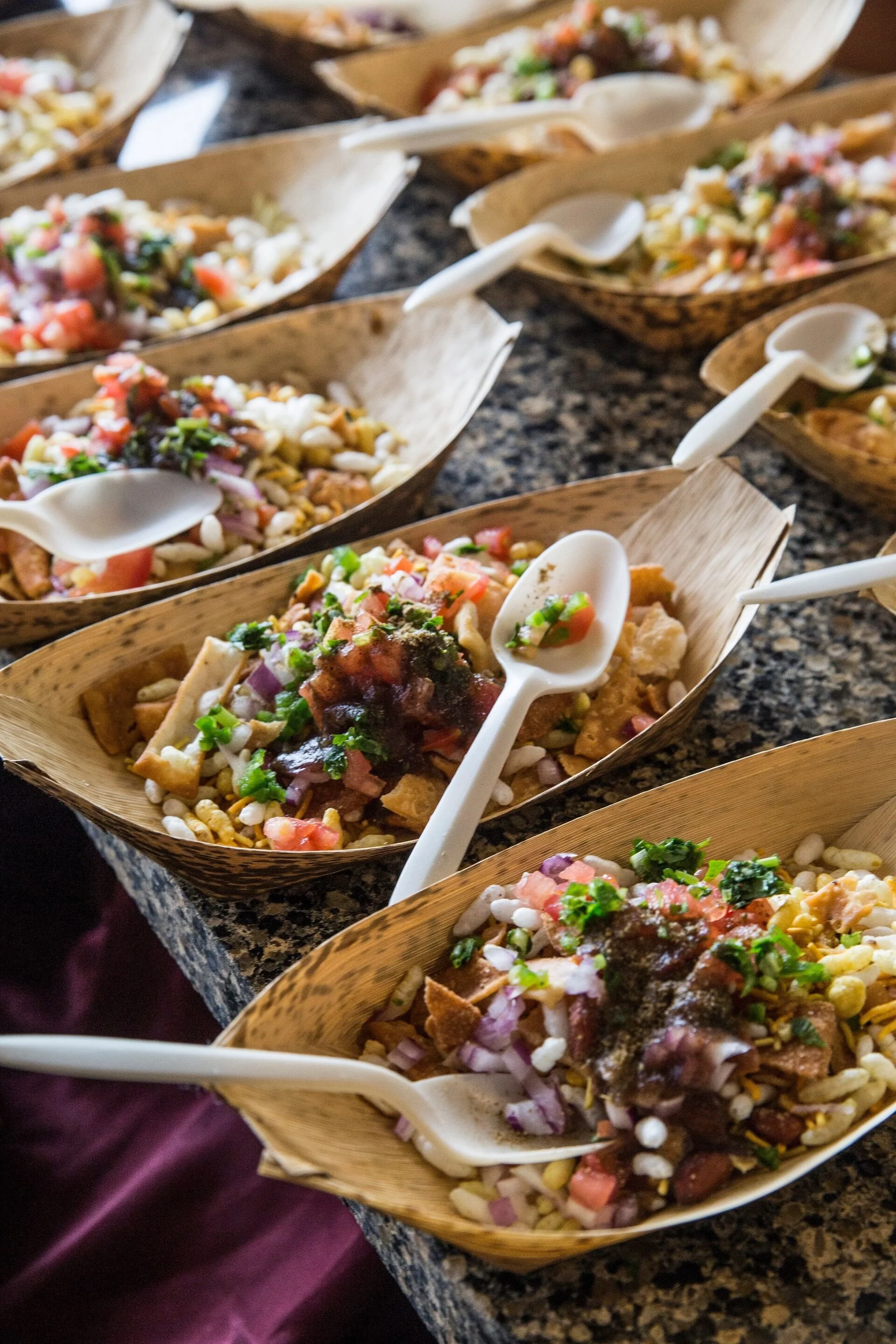 Multiple bowls of Mexican salad on a granite countertop, each with colorful ingredients like corn, tomatoes, and herbs, served with white plastic spoons.