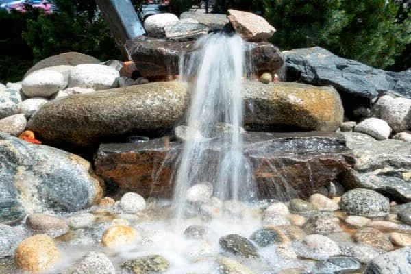 Small waterfall with water flowing over rocks and surrounded by various stones and greenery.