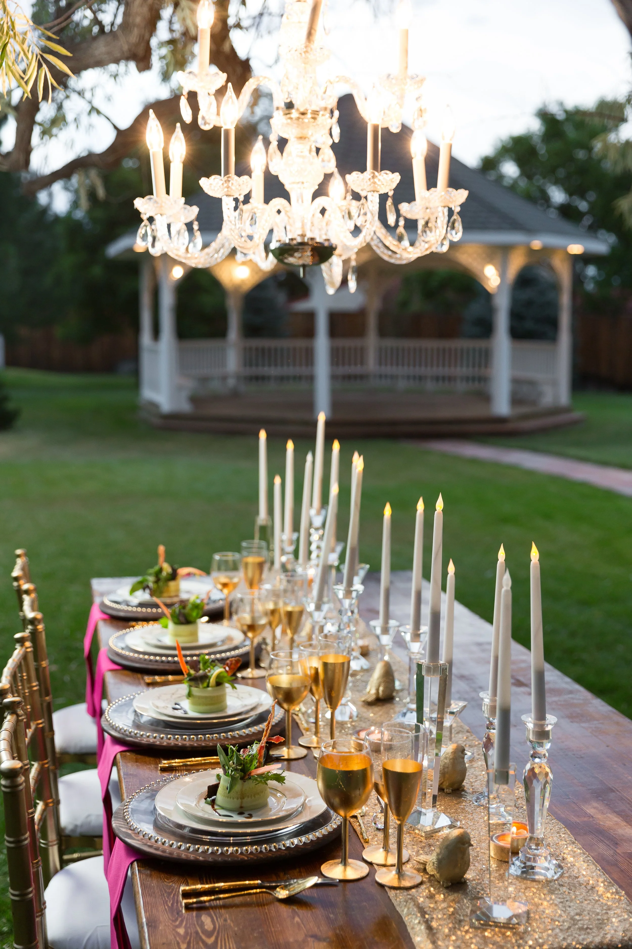 An outdoor dinner party setup with a long wooden table decorated with tall white candles in crystal holders, glassware, and elegant place settings. A crystal chandelier hangs above, and there is a gazebo in the background surrounded by greenery.