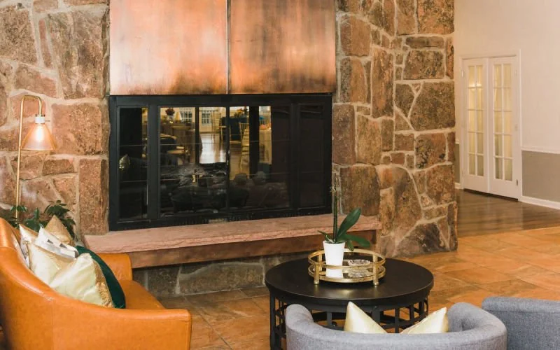 Living room with a large stone fireplace, a black coffee table with a plant in a white pot, an orange armchair with gold and dark green pillows, and a gray armchair with light-colored pillows. There is a wooden floor and glass-paneled double doors.