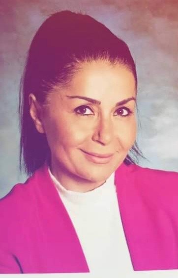 Porträt einer Frau mit dunklem, hochgestecktem Haar, rosa Jacke und weißem Top vor einem neutralen Hintergrund.