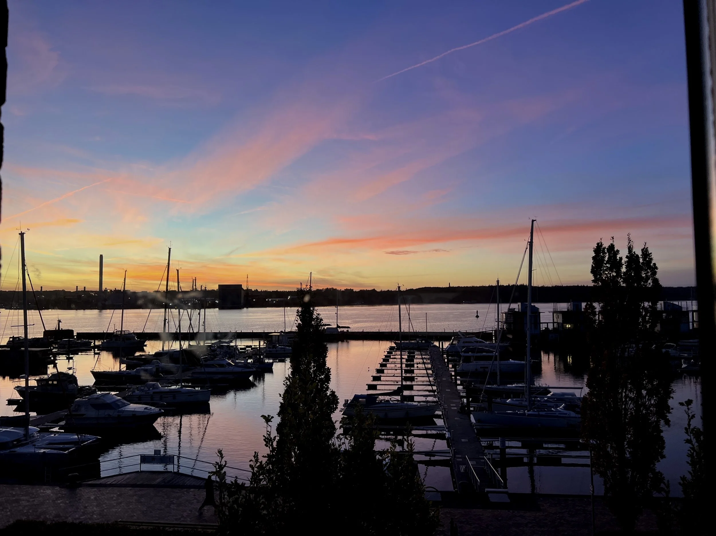 Blick auf einen Yachthafen bei Sonnenuntergang mit reflektierendem Wasser und einem farbenfrohen Himmel in Pink, Orange und Blau, mehrere Segelyachten und Bootsstegen.