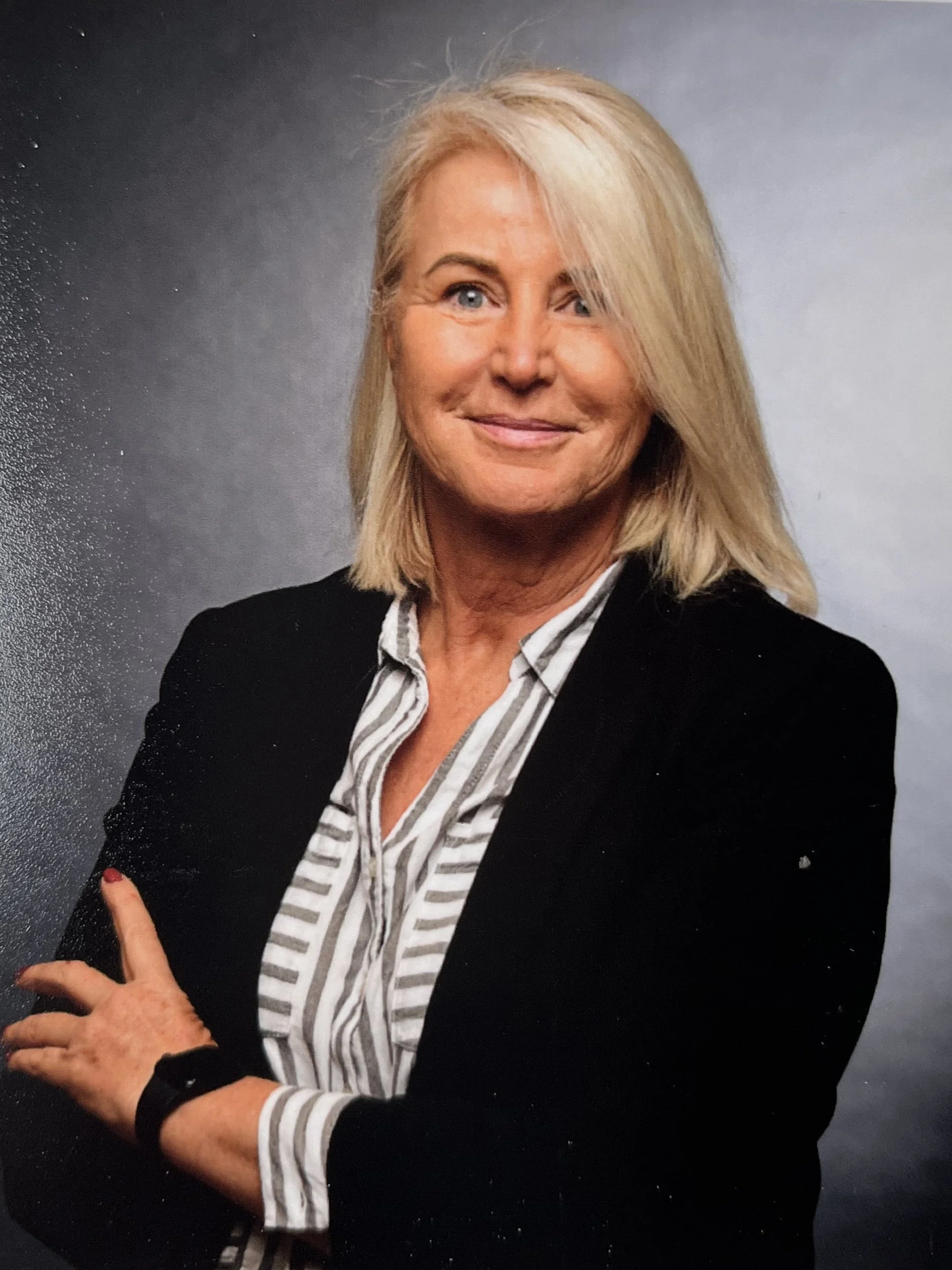 Porträt einer älteren Frau mit blondem Haar, die einen schwarzen Blazer und ein gestreiftes Hemd trägt, vor hellem grauem Hintergrund.