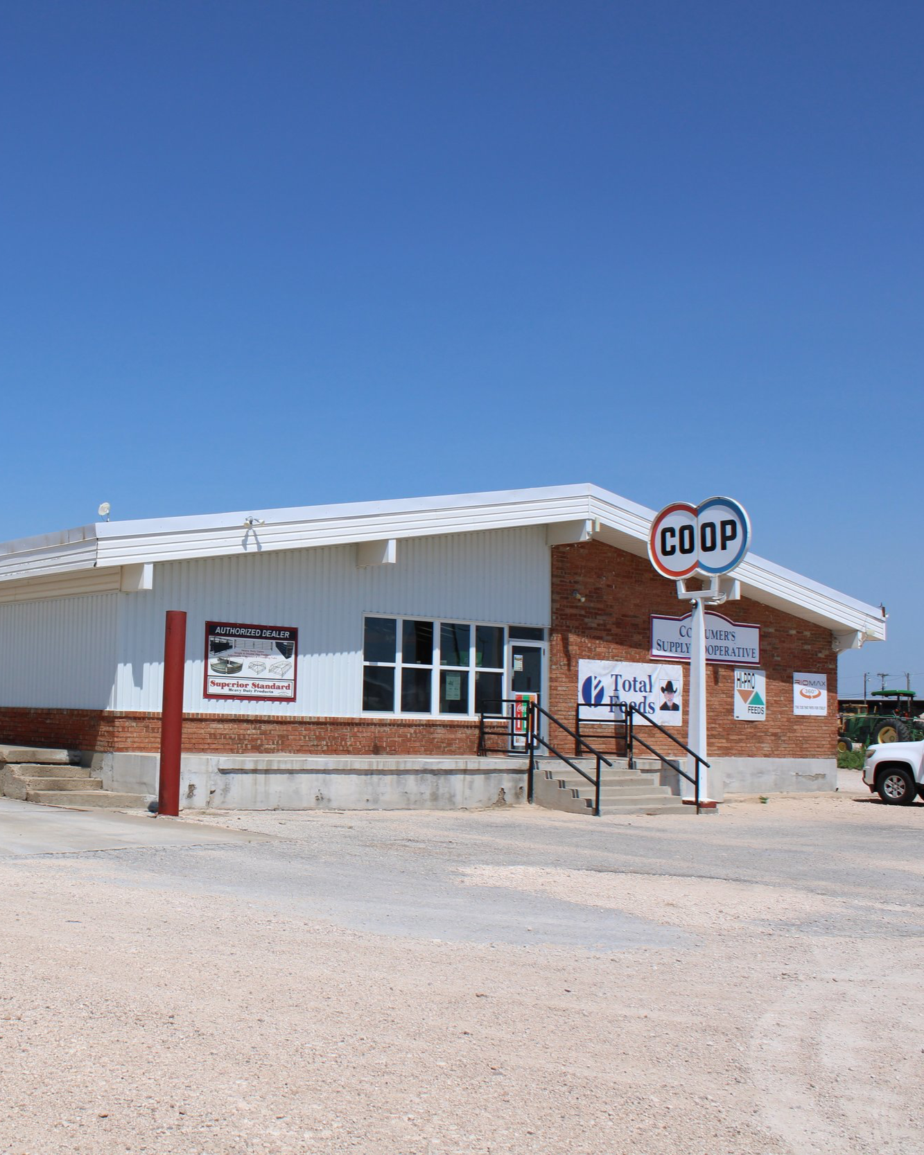 A small retail store with a white metal roof and brick walls, featuring a CO-OP sign, outside stairs, and various advertisements for products including Total Foods and Feeds.